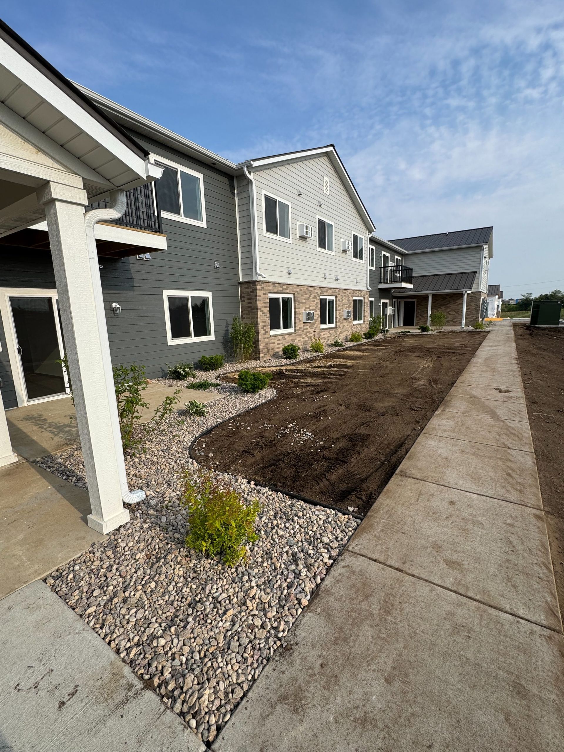 A multi-story apartment building with grey siding and stone accents, featuring a landscaped yard and concrete sidewalk.