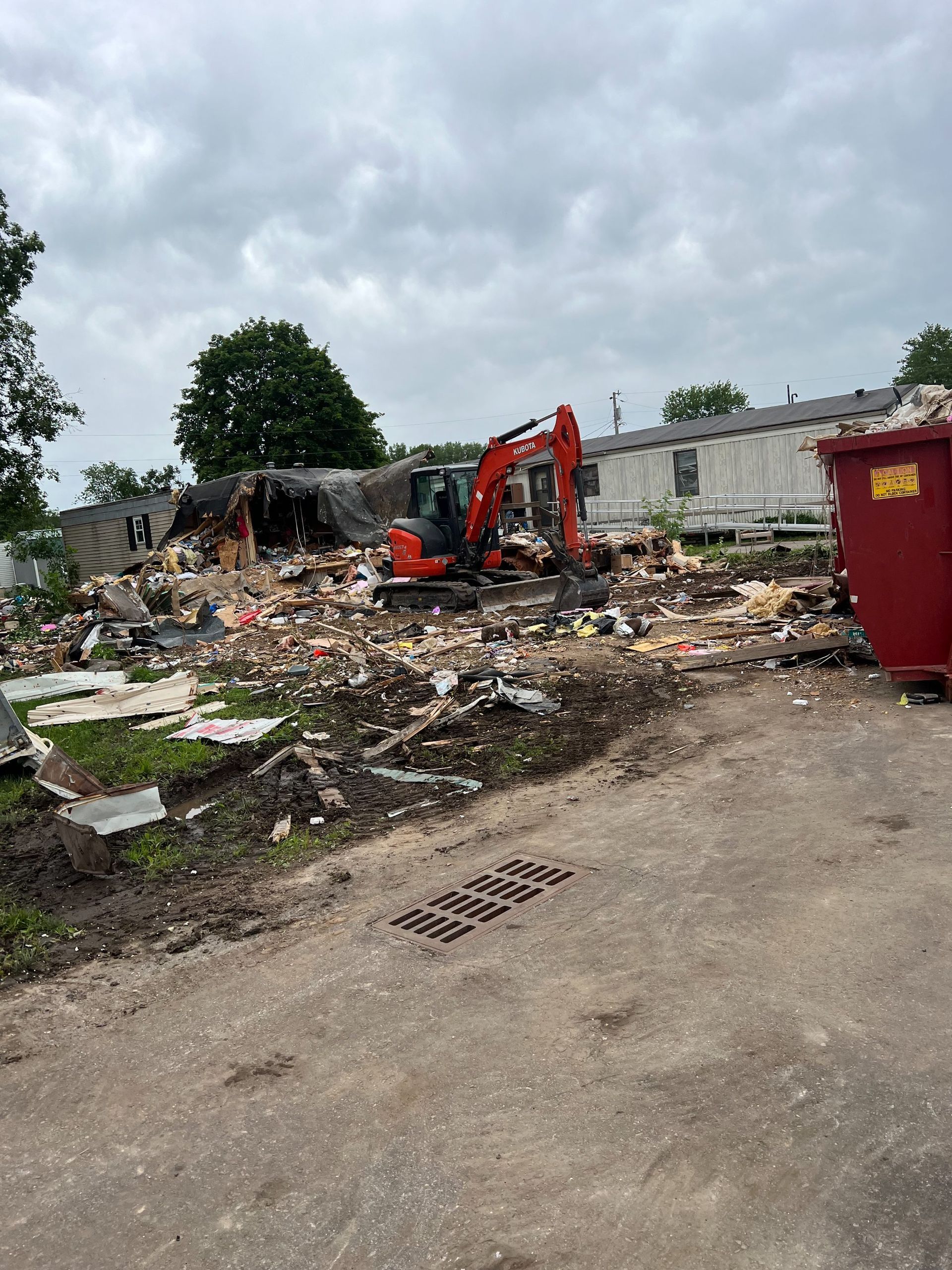 An orange excavator at a demolition site surrounded by building debris with a red dumpster nearby.