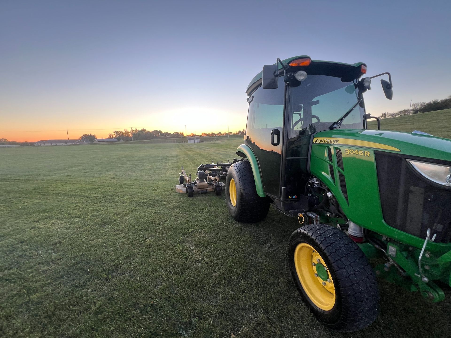 A green John Deere tractor mows a large field at sunrise.