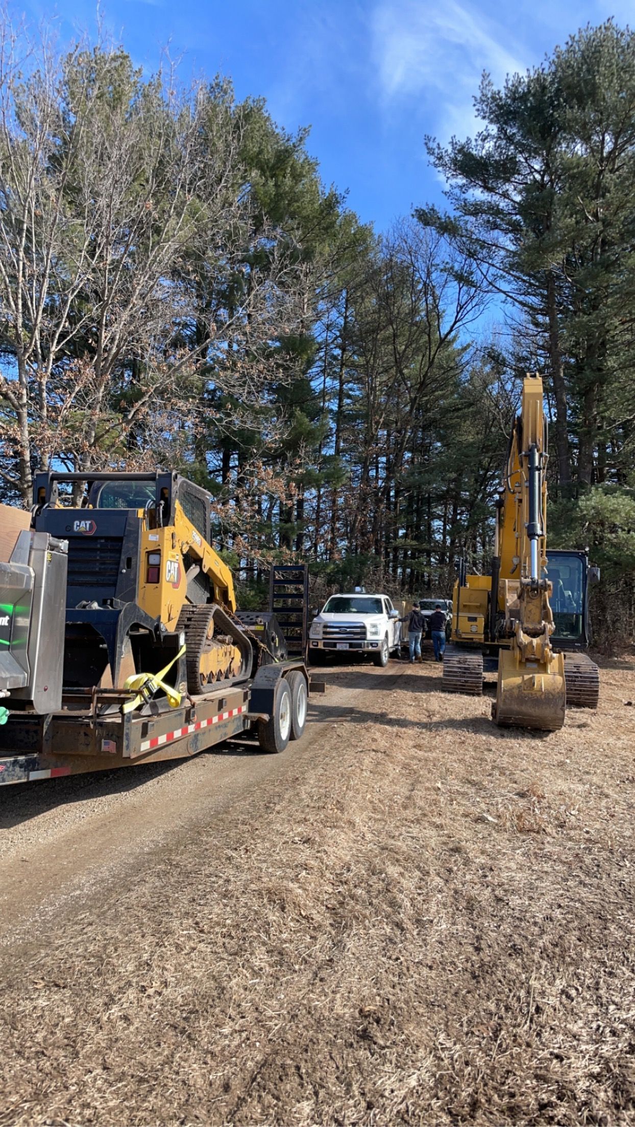 A yellow skid steer on a trailer parked next to a yellow excavator on a dirt clearing near trees under a blue sky.