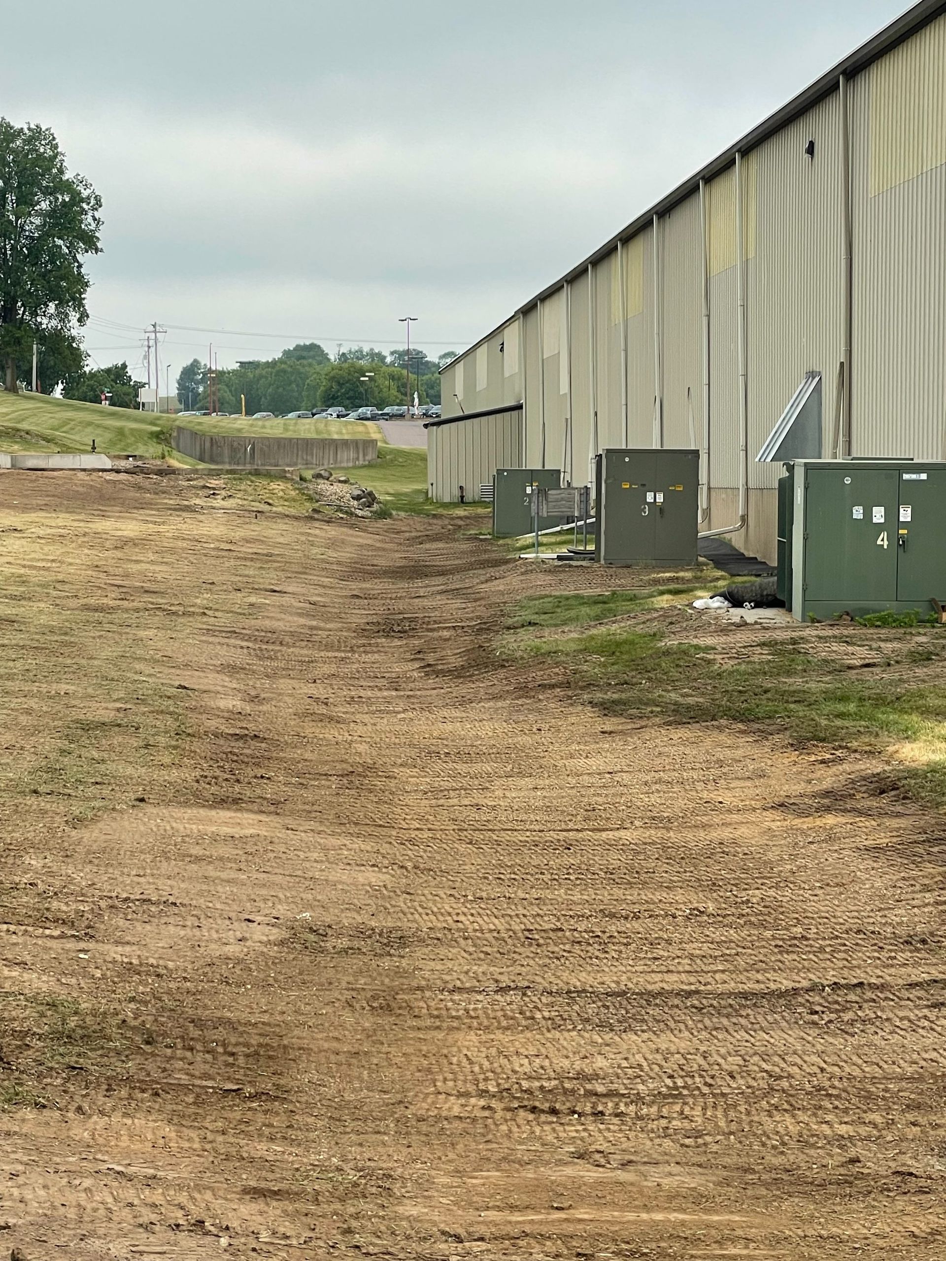 Green utility boxes sit beside a long, beige industrial building on a patch of uneven, cleared dirt ground.