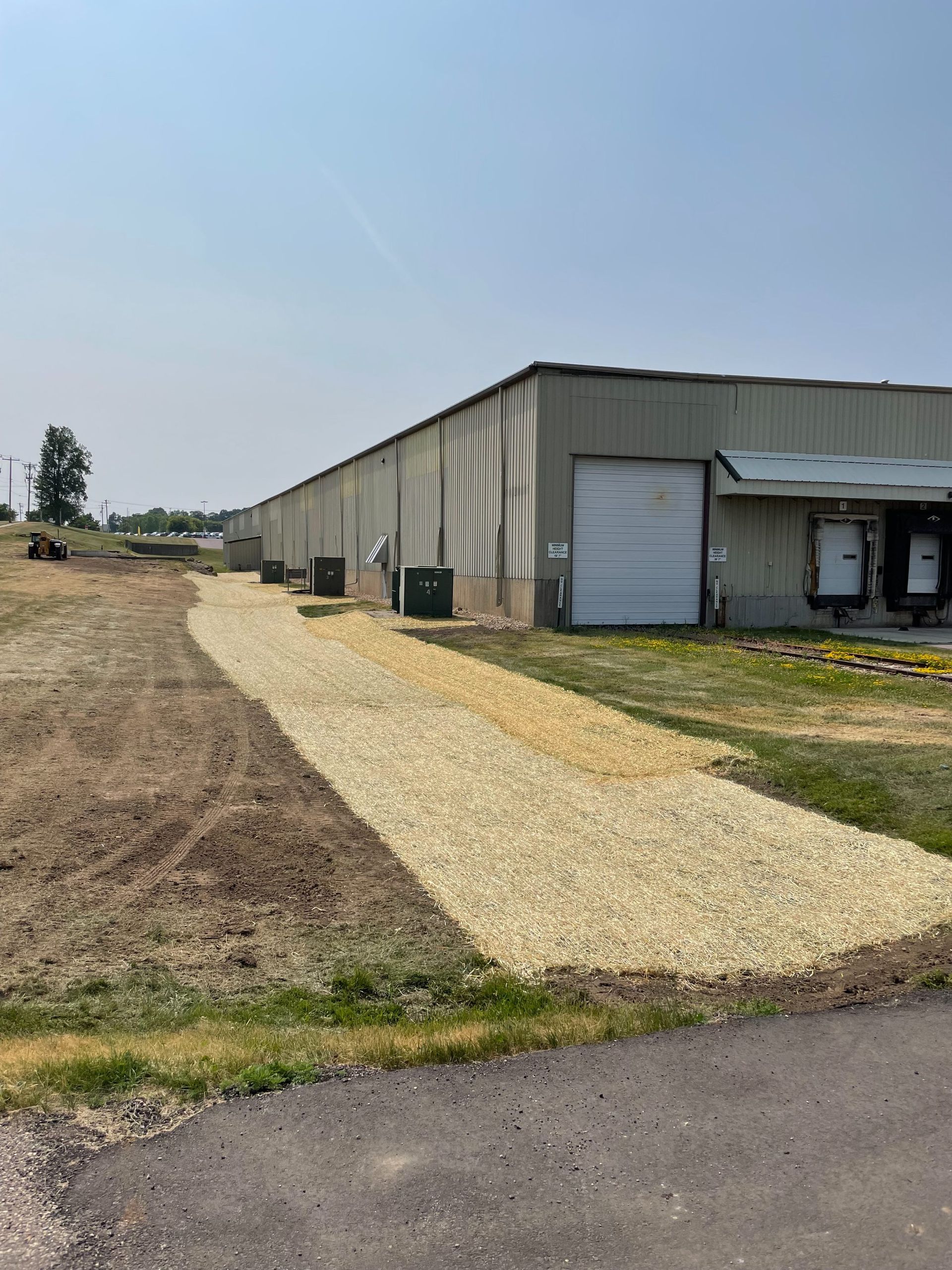 A newly laid gravel path runs alongside a long metal warehouse building on a sunny day.