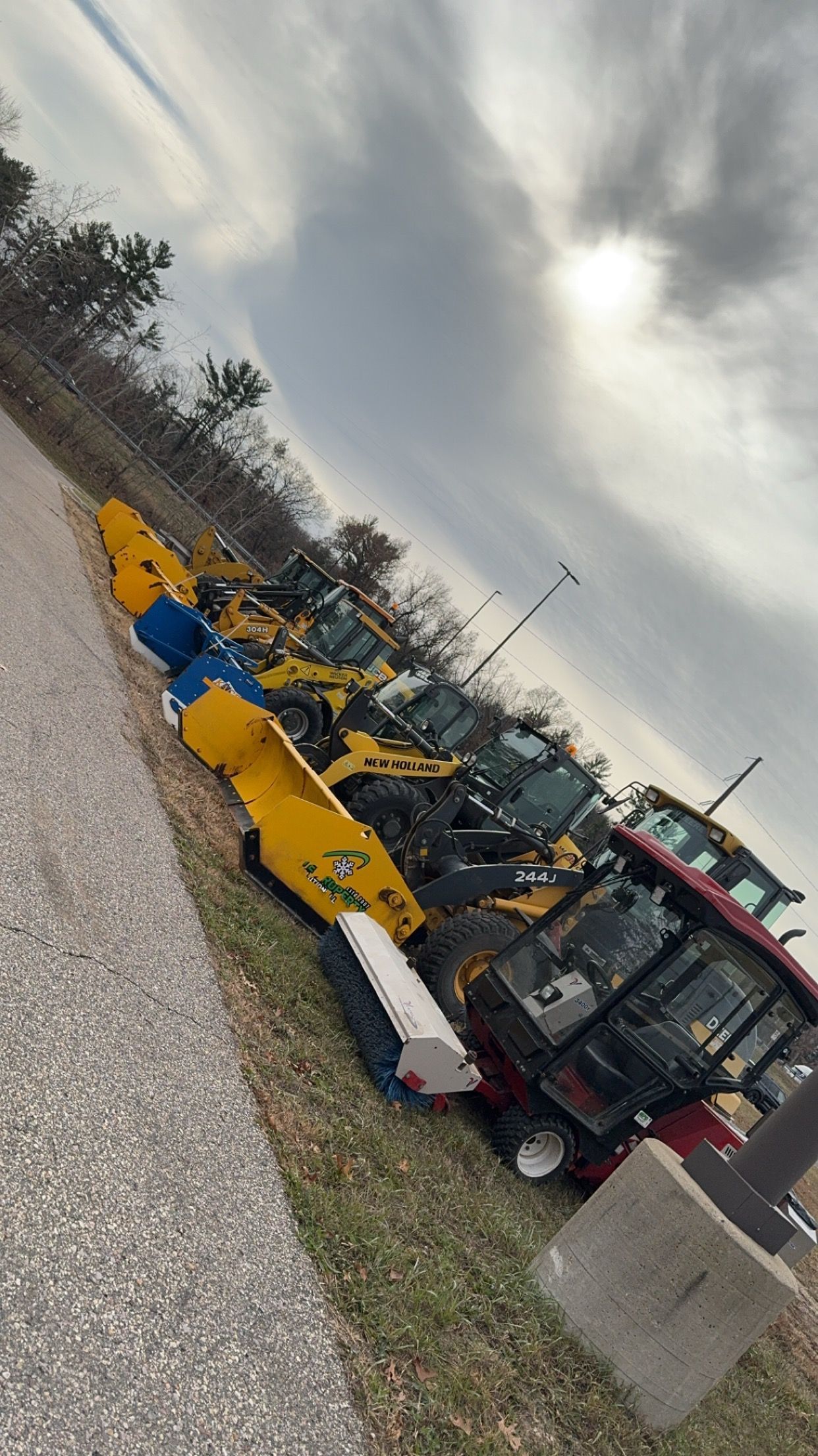 A line of yellow construction vehicles and heavy machinery parked on a gravel lot under a cloudy, overcast sky.