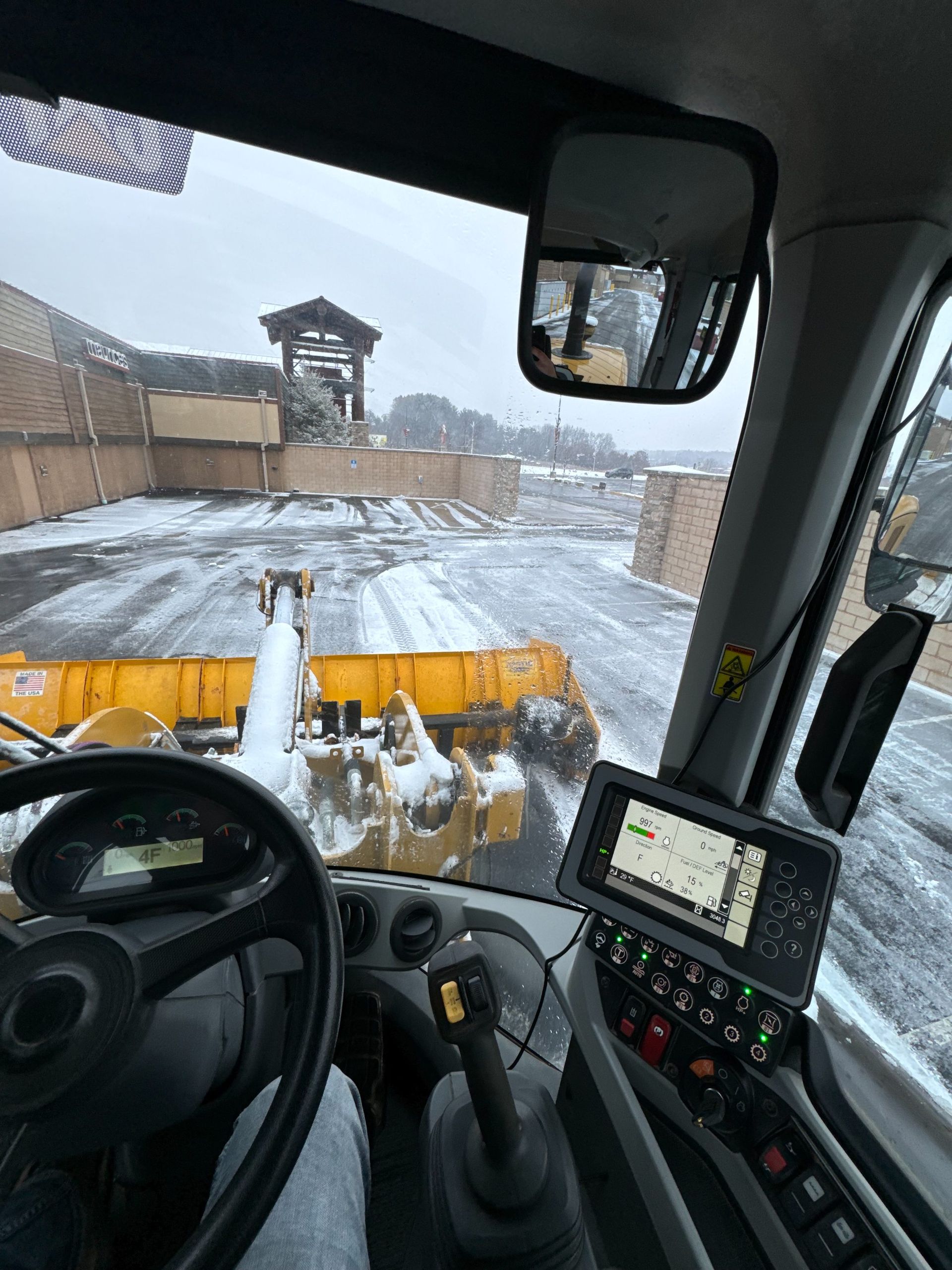View from inside the cab of a yellow snowplow clearing a snowy parking lot near a building.