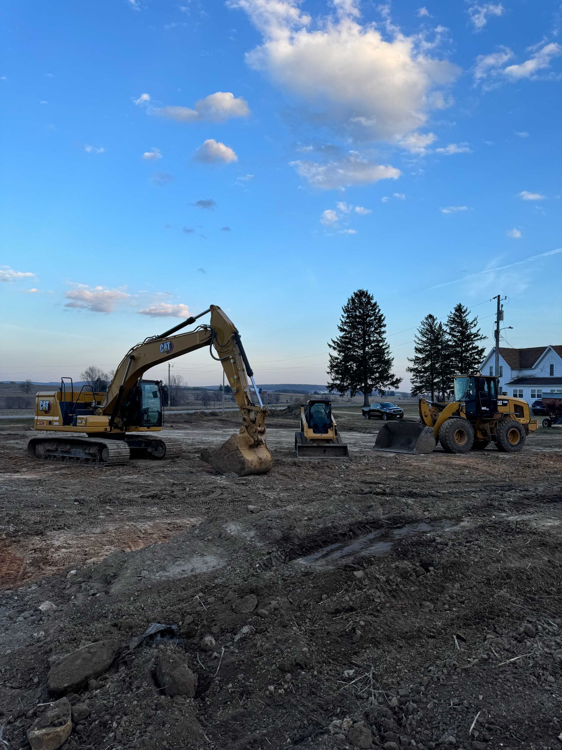 A yellow excavator, a skid steer, and a wheel loader sit on a dirt construction site under a blue sky.