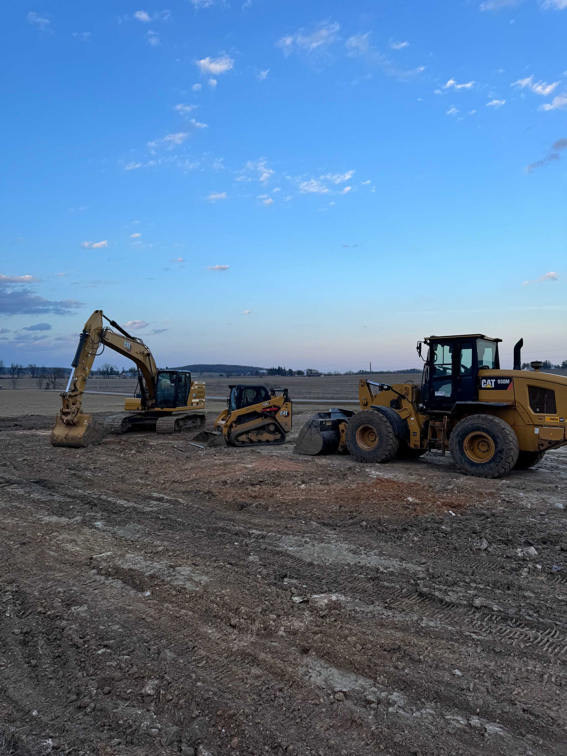 A yellow excavator, a small tracked loader, and a large front-end loader parked on a rocky construction site at dusk.