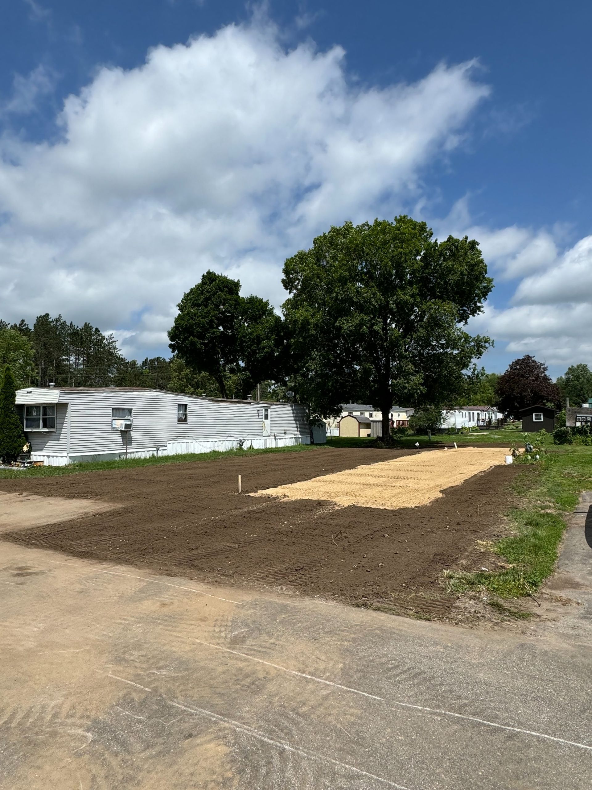 A freshly tilled, rectangular garden plot with a straw-covered section next to a mobile home under a blue sky.