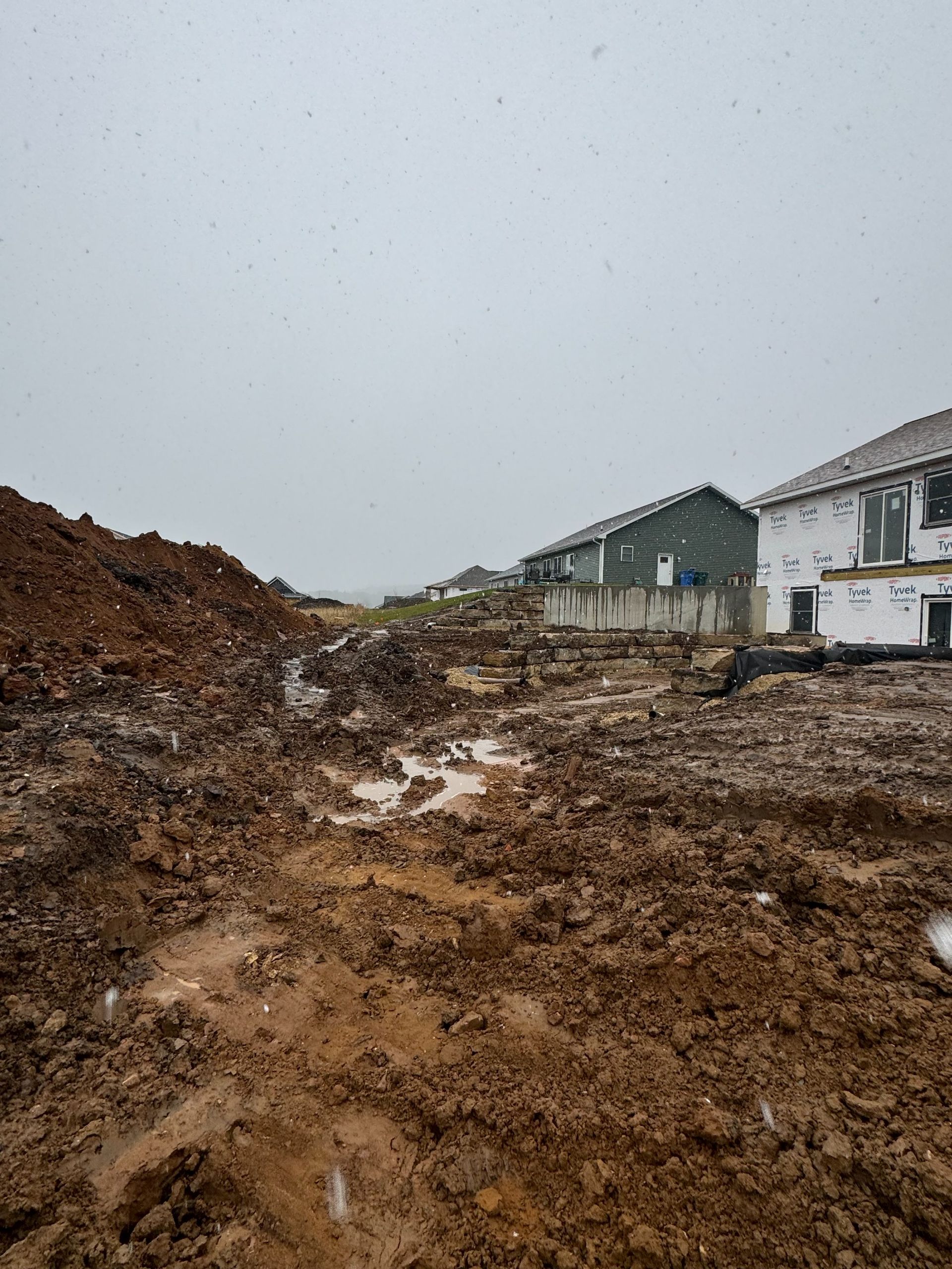 A muddy, undeveloped construction site with a large dirt pile and a house under construction in the background.
