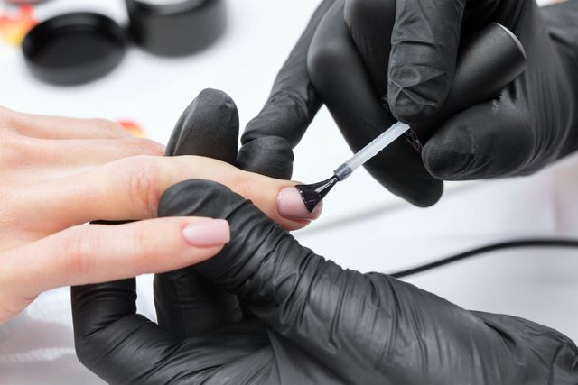 Manicure che applica lo smalto sull'unghia di una cliente, indossando guanti neri.
