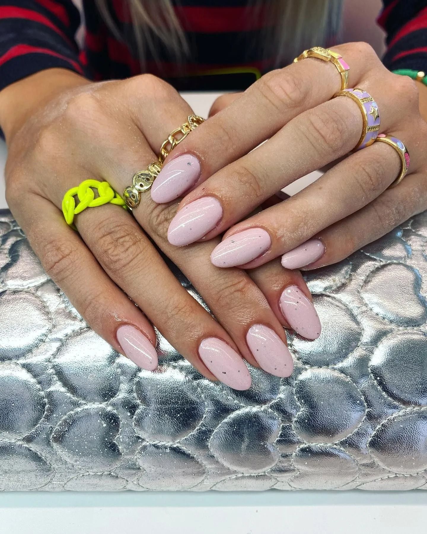 Mani con unghie ovali color rosa chiaro e anelli dorati su una superficie argentata.