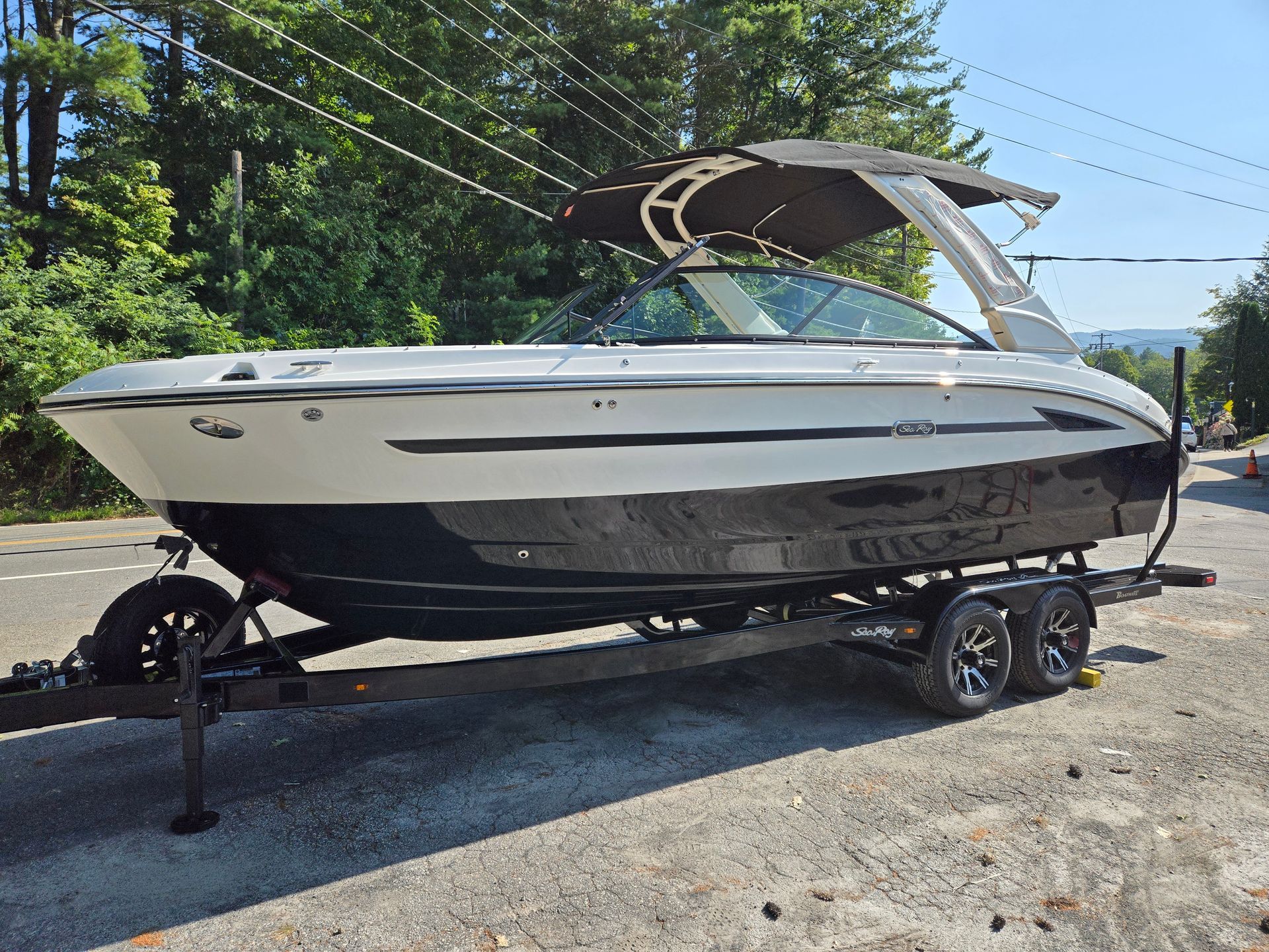 White and black motorboat on a trailer, parked on a road with trees in the background.