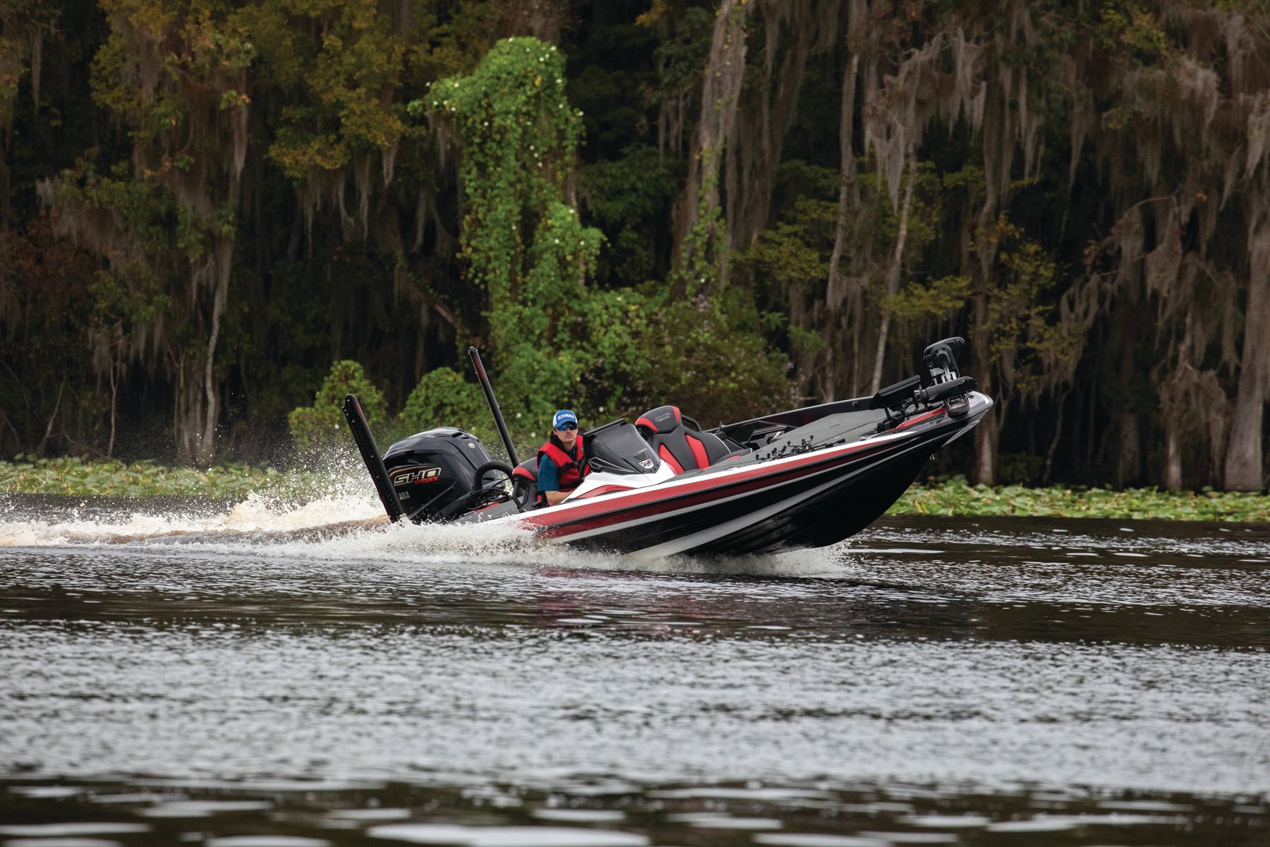 Bass boat speeding through murky water near moss-covered trees.