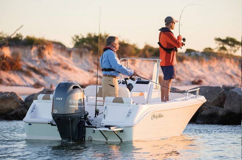 Two men fishing from a white boat near a sandy shore. One steers, the other casts a line.