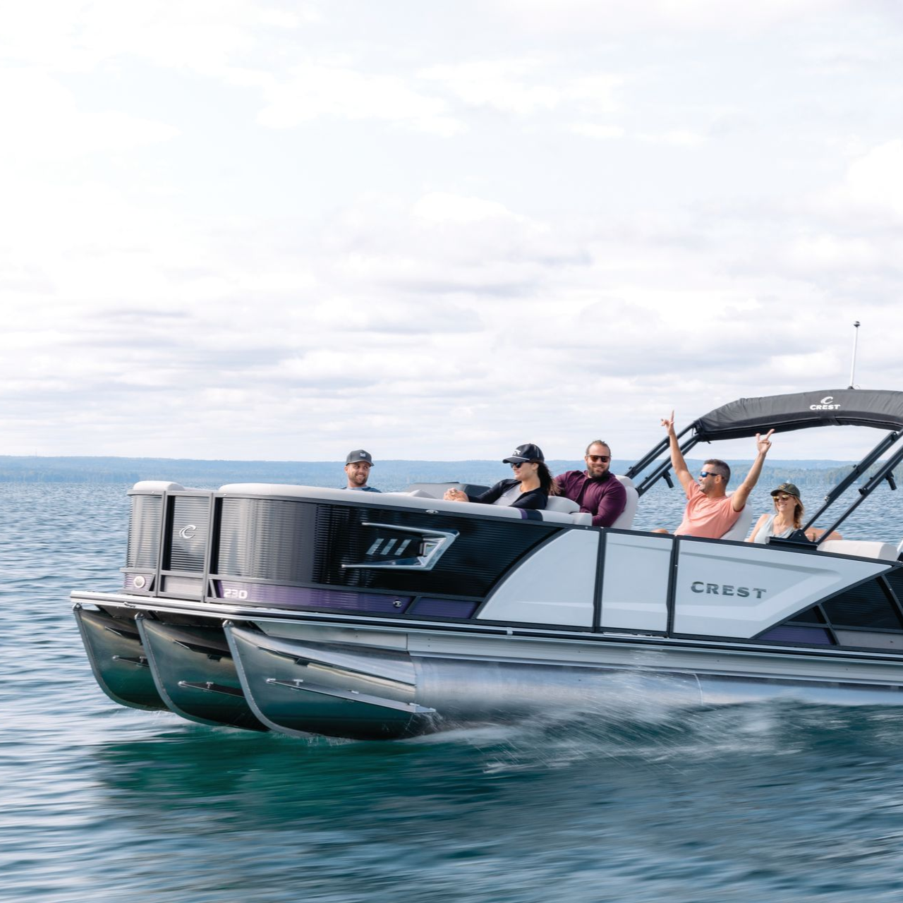 Pontoon boat with five people on a lake, waving, under a cloudy sky.