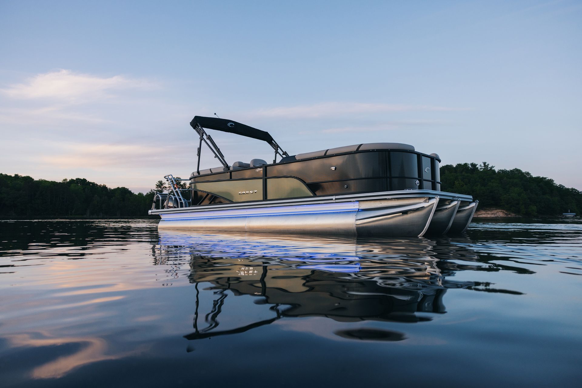 Pontoon boat floating on water, illuminated by blue lights, with trees in the background at dusk.