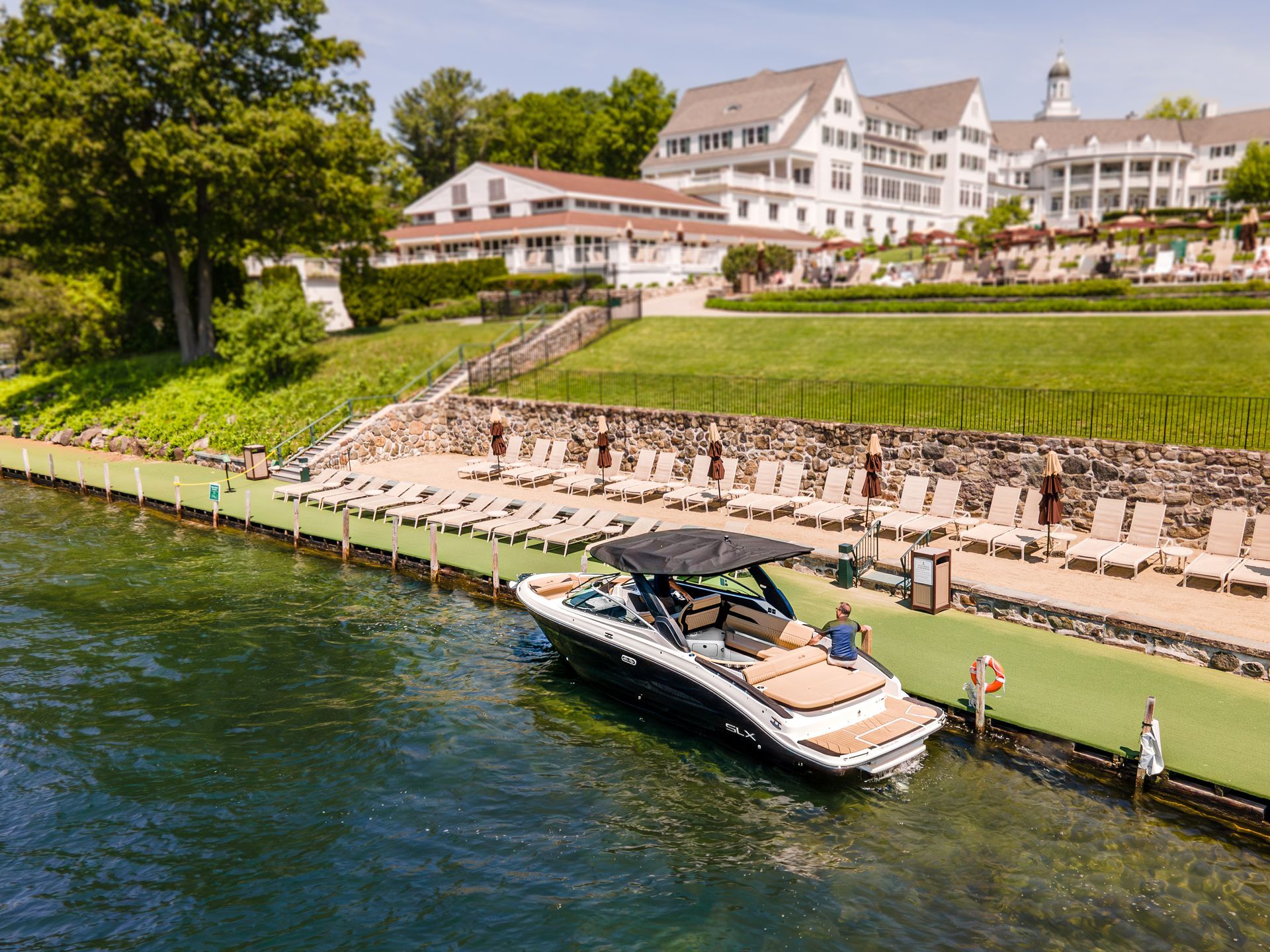 Boat docked by a grassy waterfront resort with white buildings and a tree-lined shore
