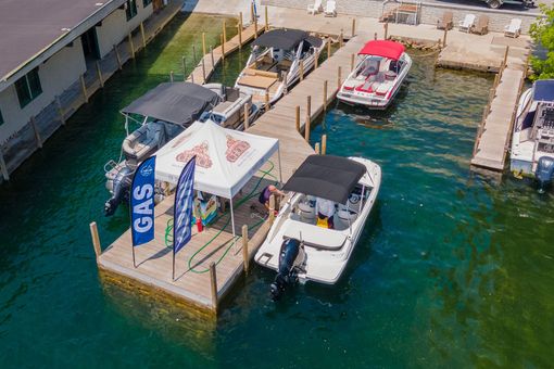 Boats docked at a pier, with a tent and flags. Aqua water and a building are visible in the background.