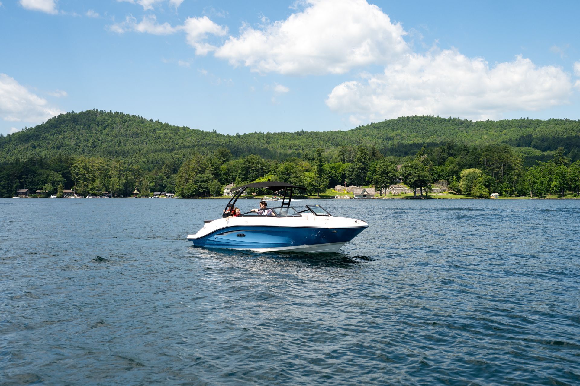 White speedboat on blue lake with people aboard, forested hills and cloudy sky in the background