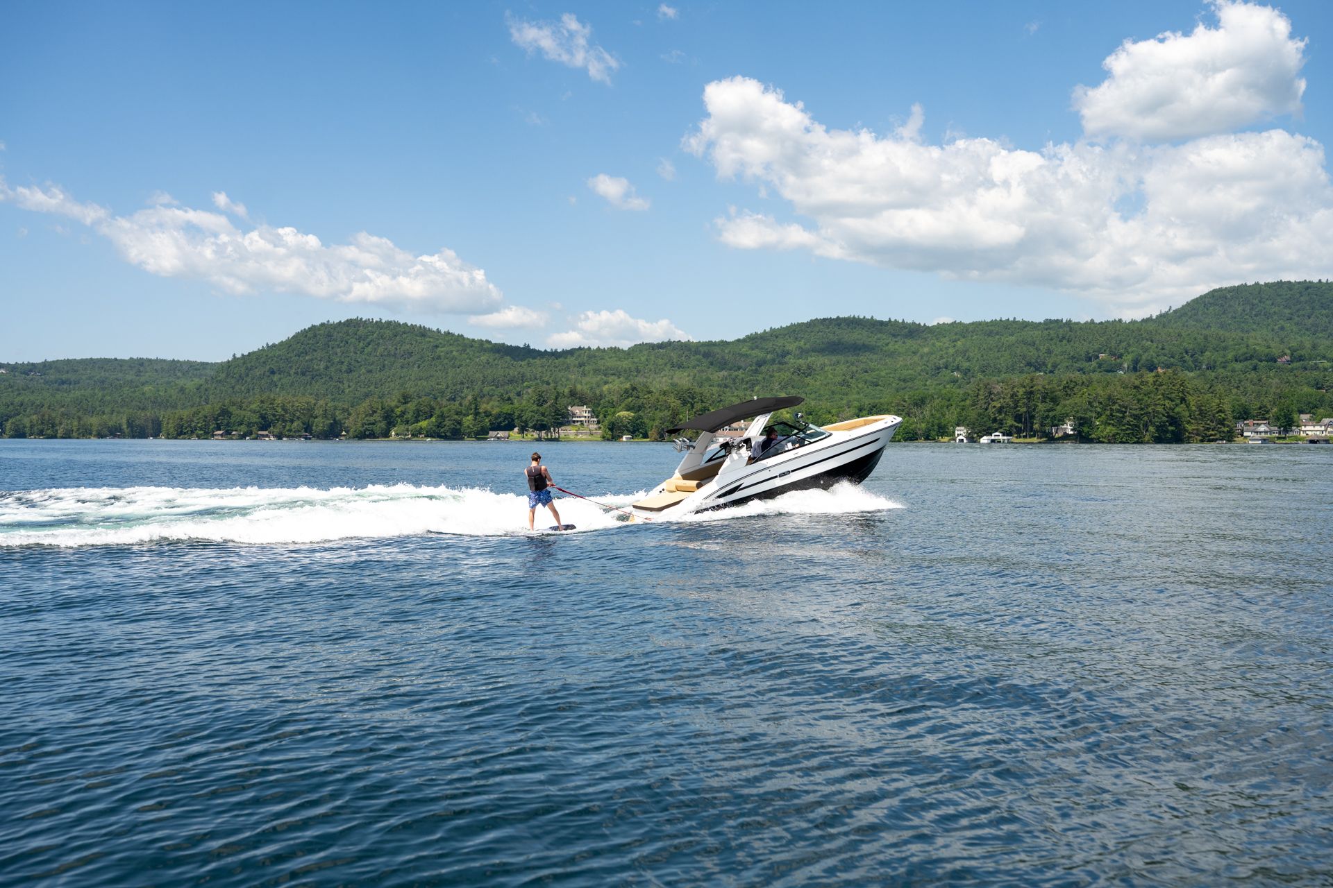 A person water skis behind a motorboat on a lake with mountains in the background. Sunny day.