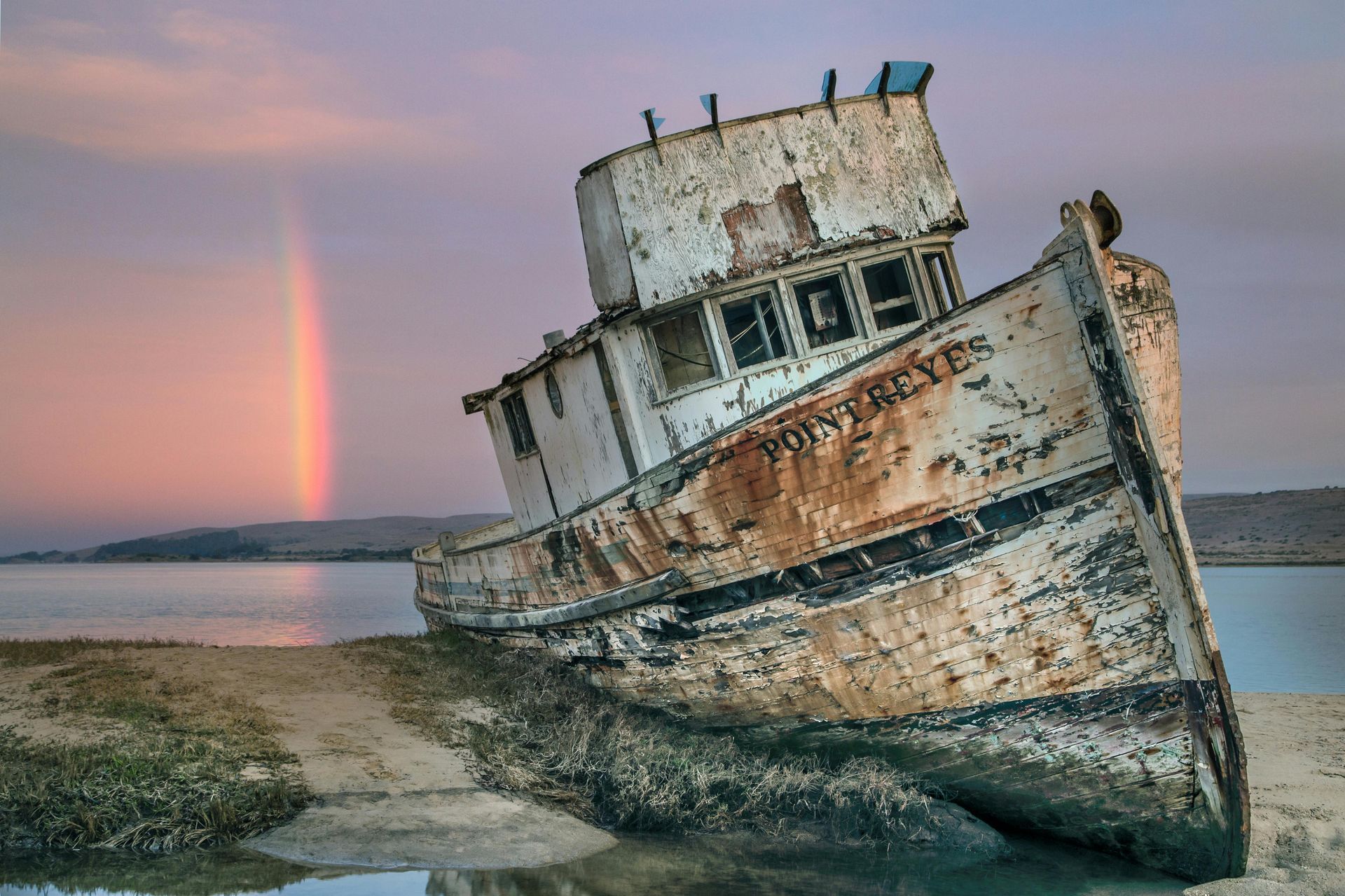 Weathered shipwreck on a beach, rainbow arching in sky.