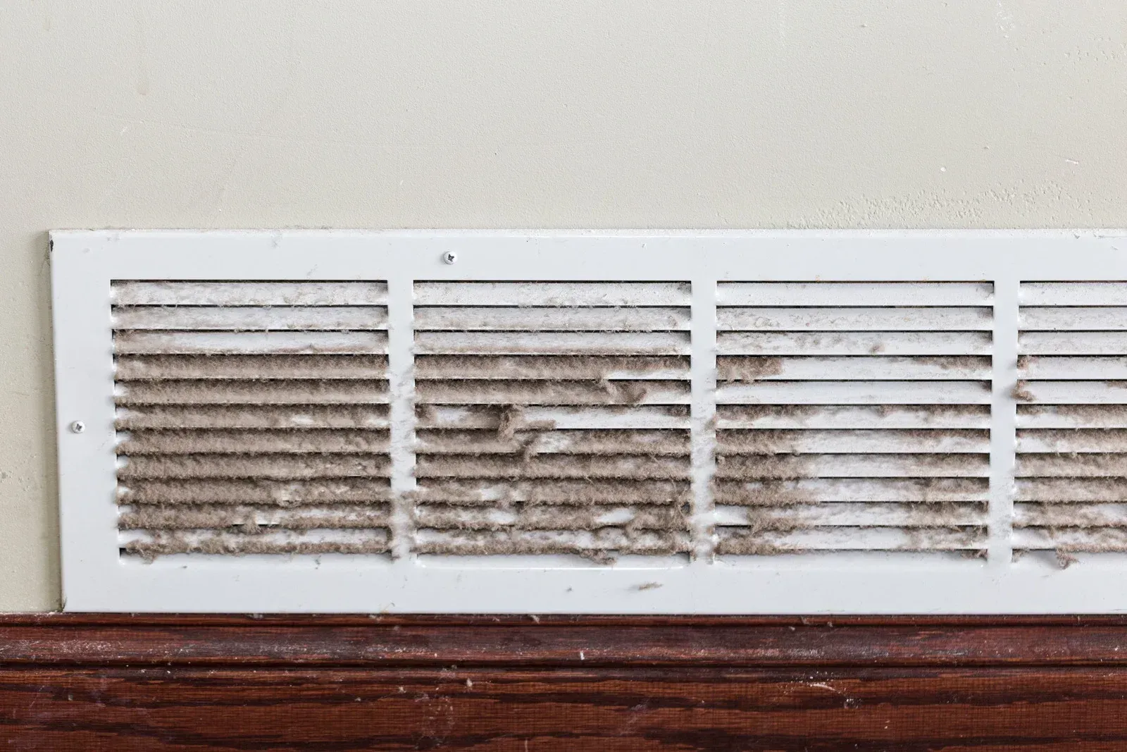 Dirty, white air vent on a beige wall above a brown wooden baseboard.