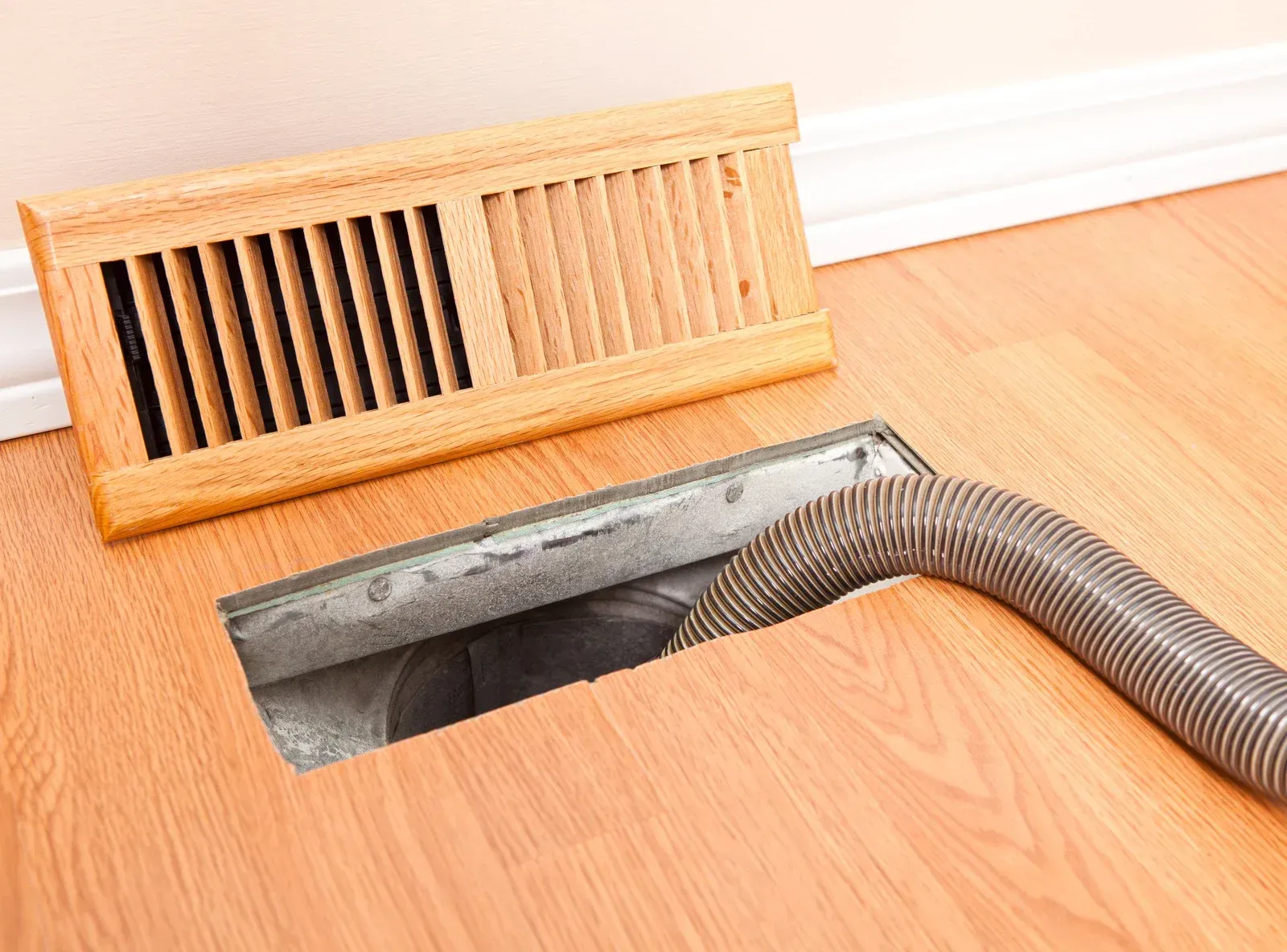 A vacuum hose cleaning a floor vent on a wood floor, wooden vent cover beside.