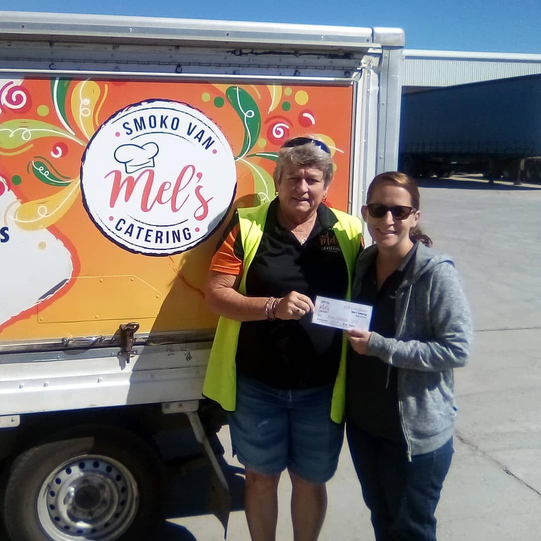 Two women holding a check in front of a catering van with a logo; daytime.— Mels Catering in Garbutt, QLD