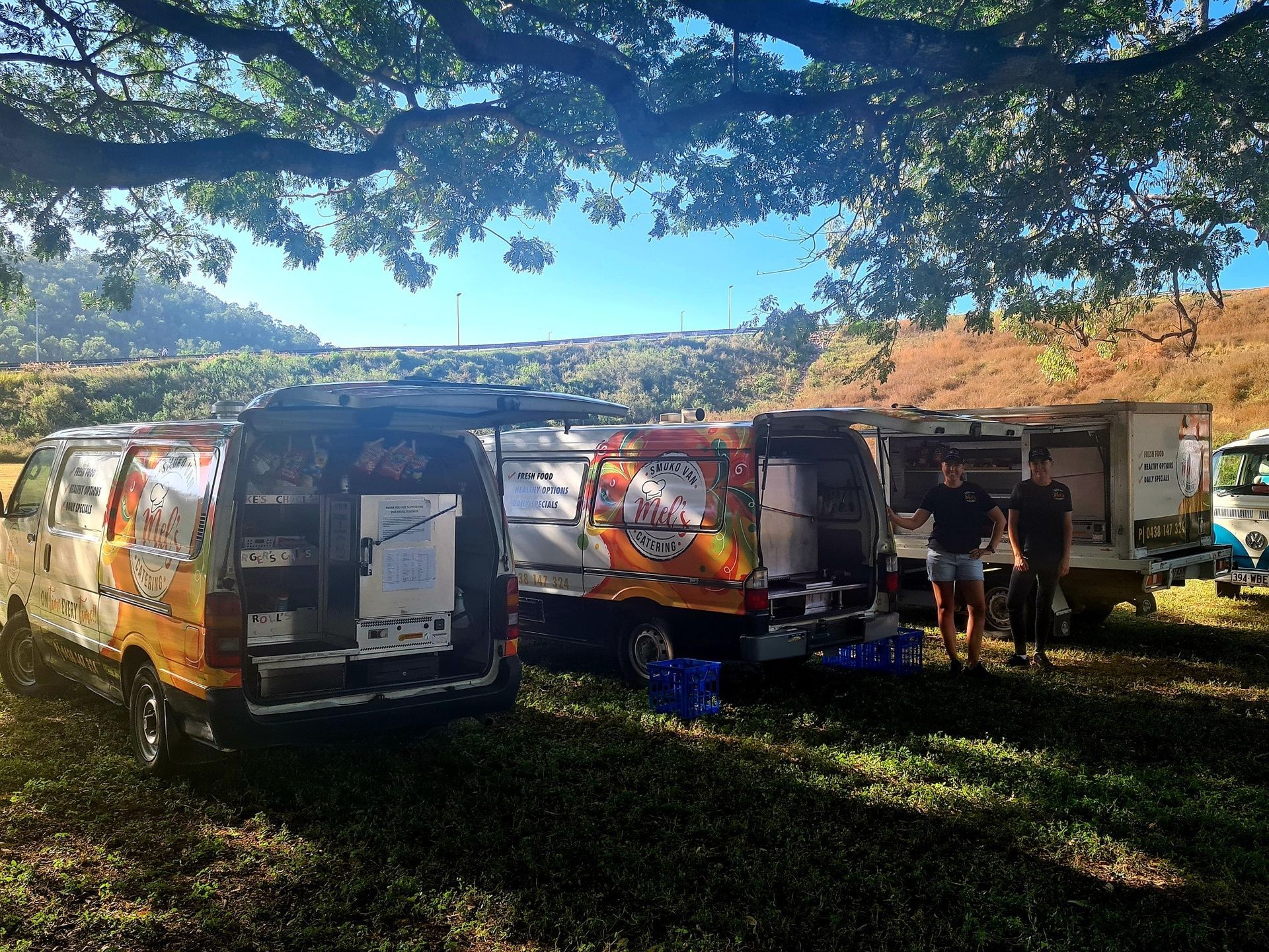 Food trucks parked under a tree, with people nearby. Sunny outdoor setting.— Mels Catering in Garbutt, QLD