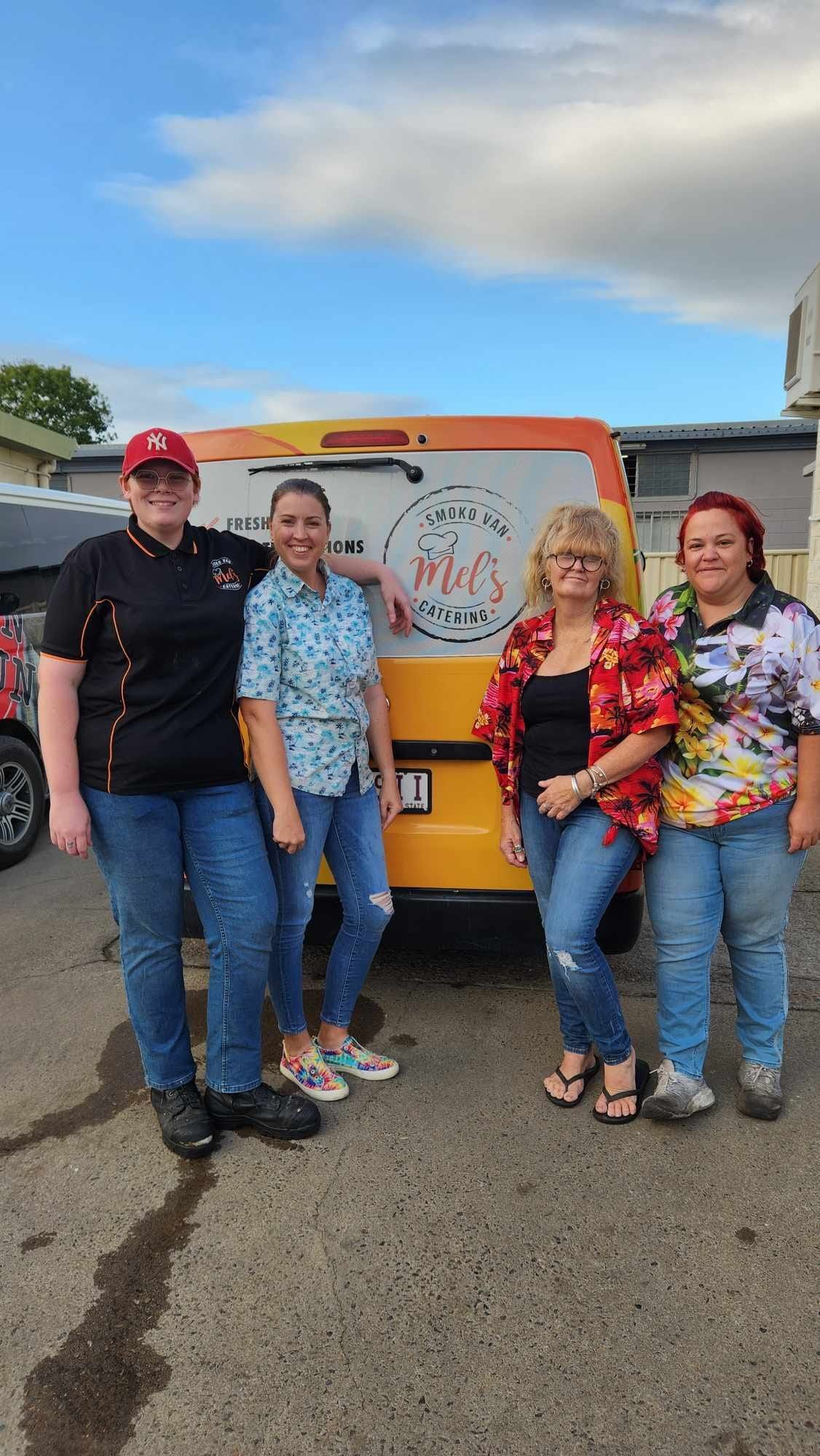 Four people stand in front of a yellow van with a logo. They wear casual clothes, smiling, under a cloudy sky— Mels Catering in Garbutt, QLD