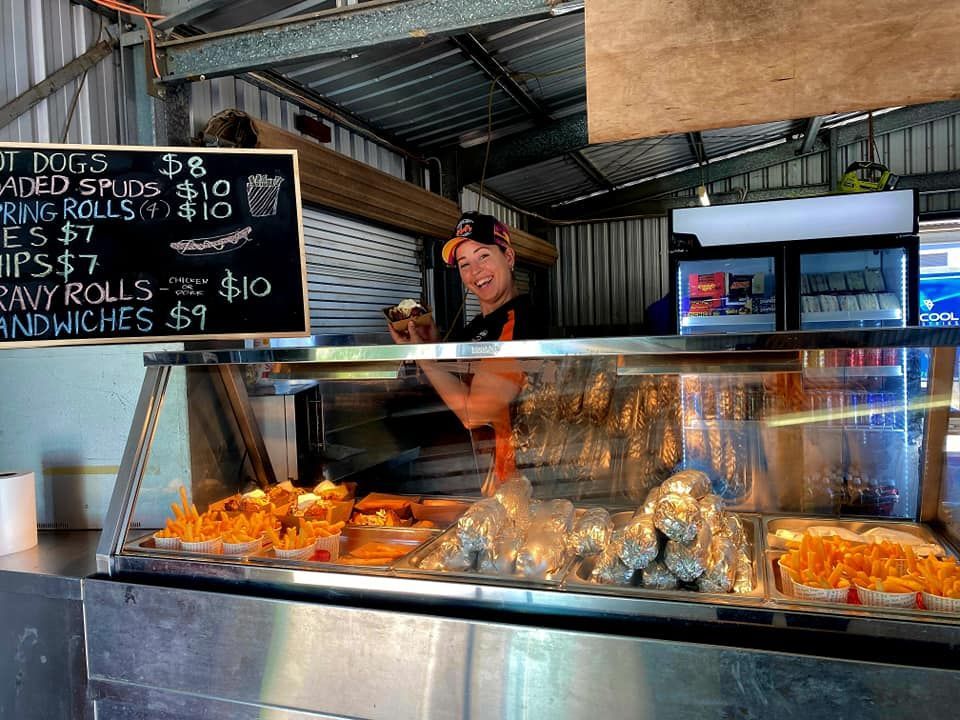 A person smiles behind a food counter, displaying hot food and a menu board.— Mels Catering in Garbutt, QLD