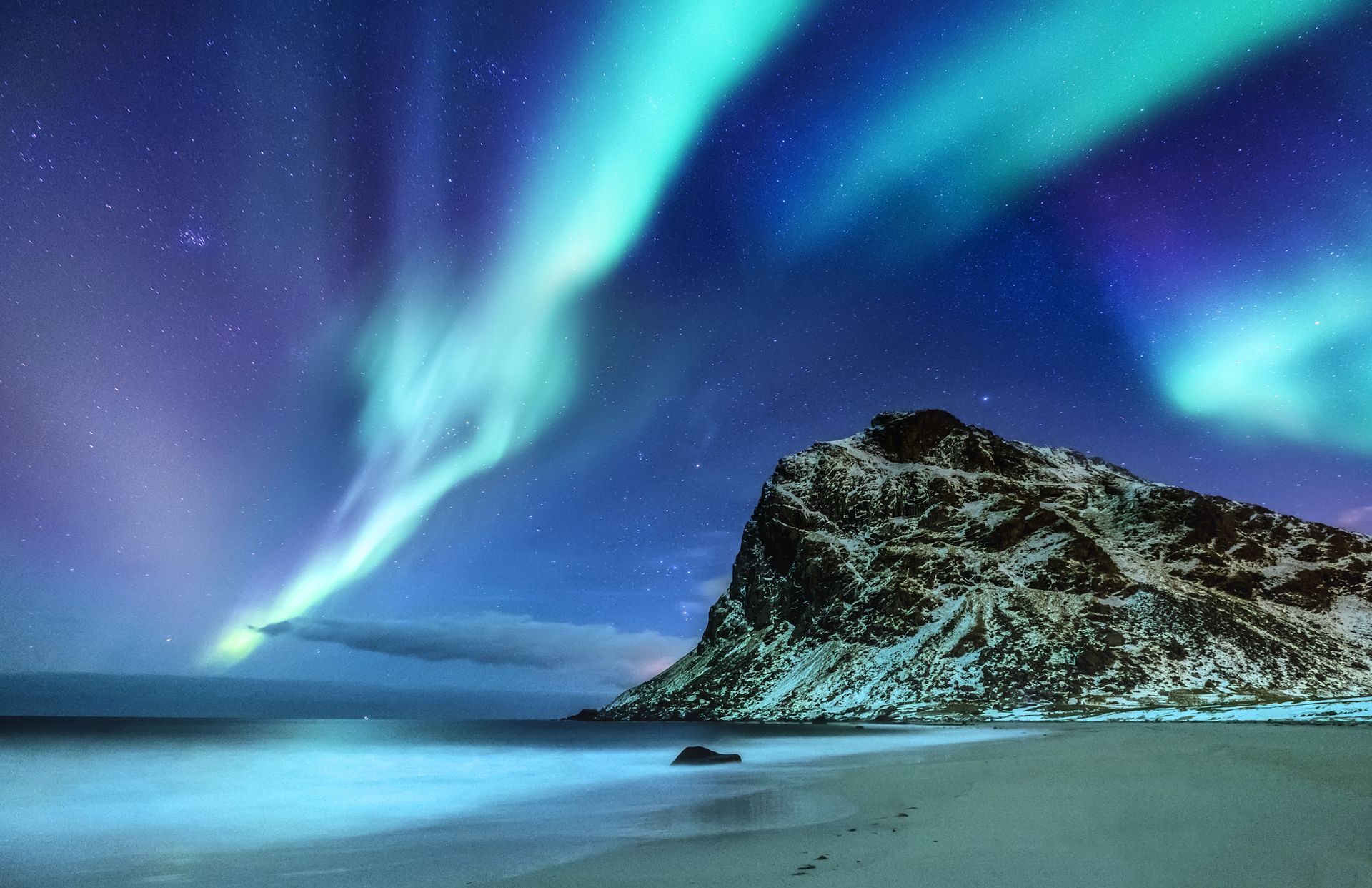 Aurora boreal sobre una montaña y una playa cubiertas de nieve, con azules y verdes vibrantes en el cielo nocturno.