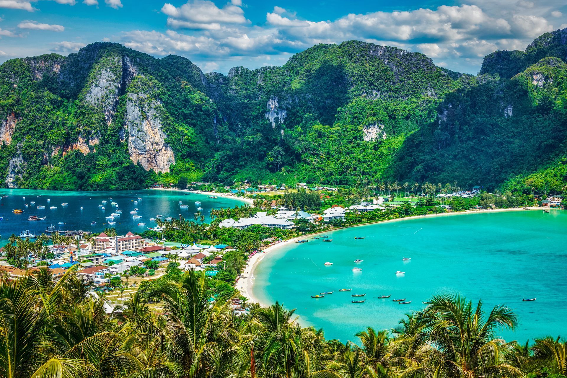 Bahía tropical con agua turquesa, exuberantes montañas verdes, barcos y una playa de arena.