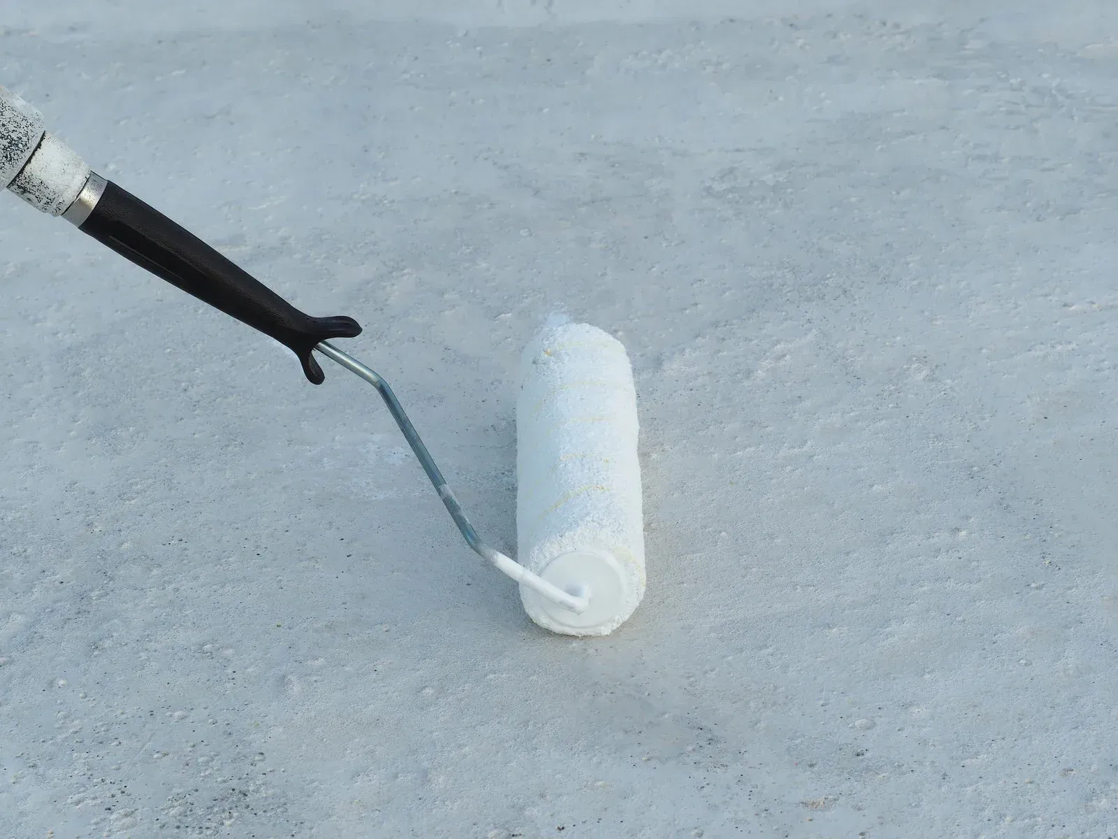 A paint roller, applying white paint to a concrete surface.