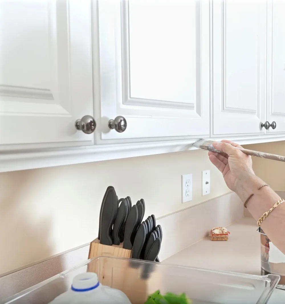 Person painting white kitchen cabinets with a paintbrush.