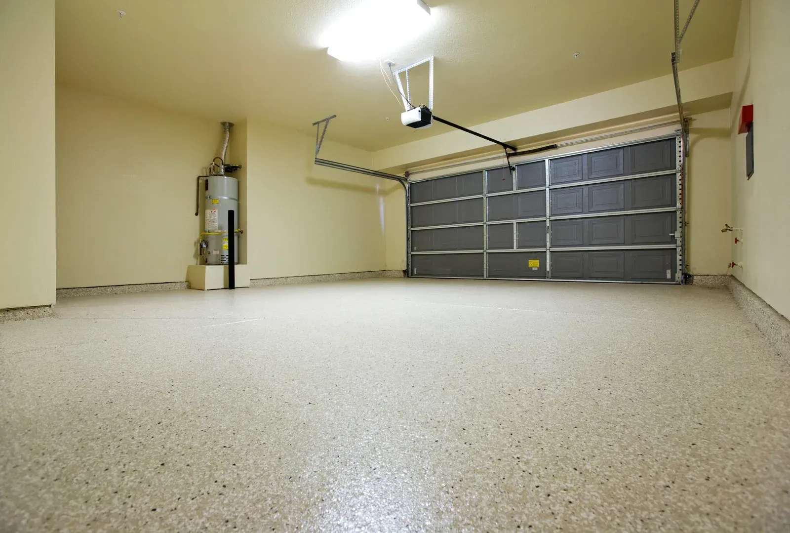 Empty garage with speckled epoxy floor, closed gray door, water heater, and overhead light.