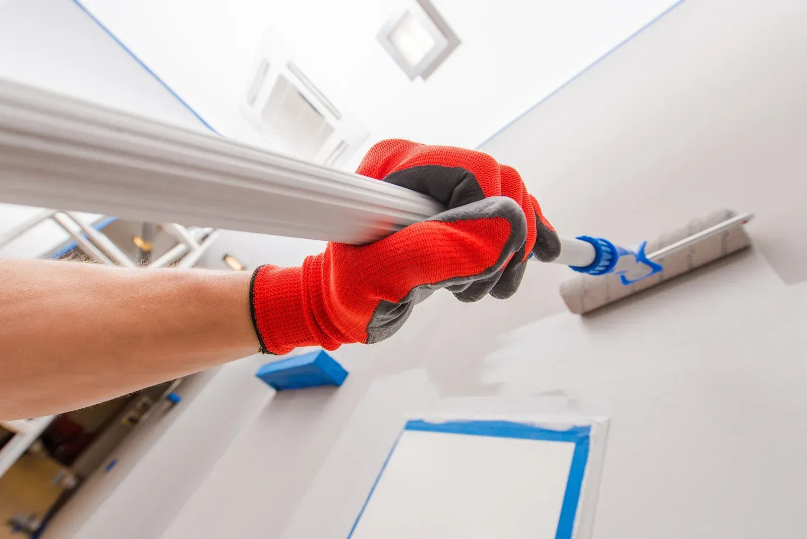 Person in red gloves painting a white wall with a roller and extension pole.