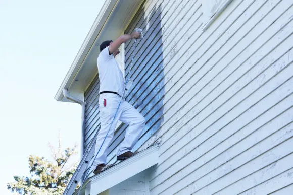 Person paints a white house siding while standing on a ladder.