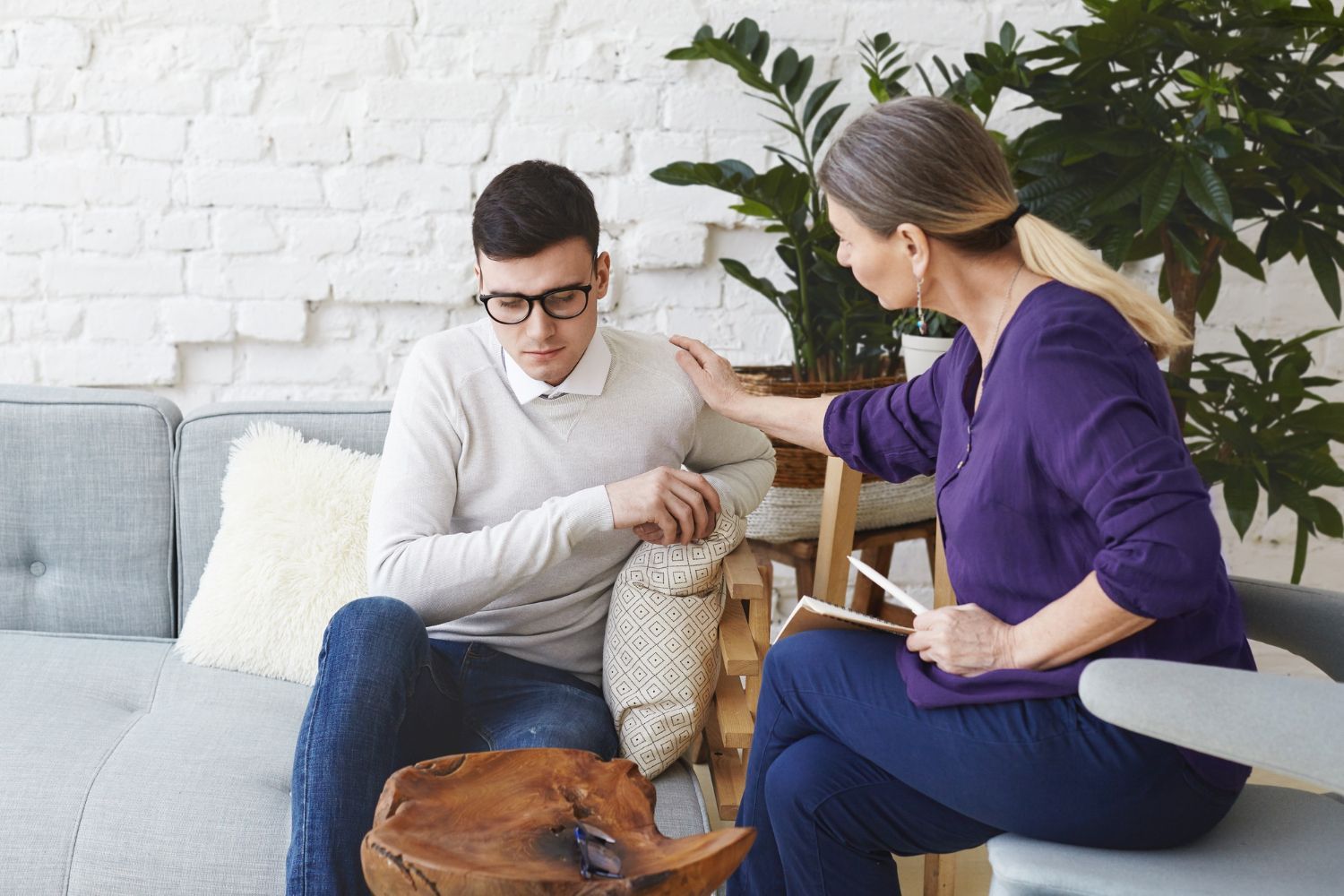Therapist comforts a person on a couch. The therapist touches the person's arm and holds a notepad.