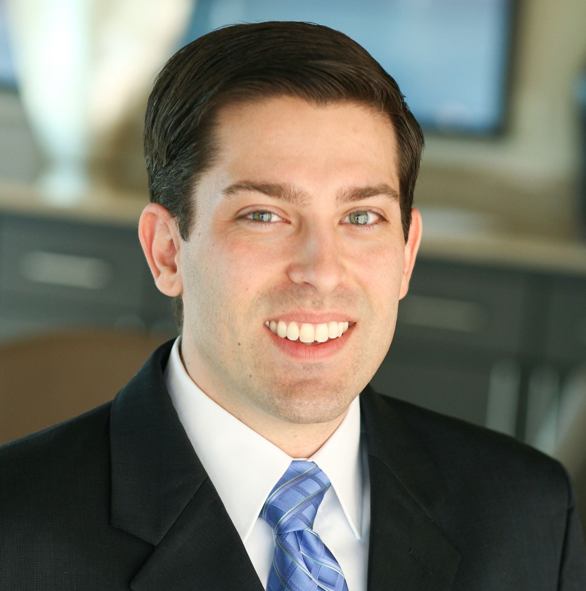 Man in suit and tie smiles at the camera; blurred office background.