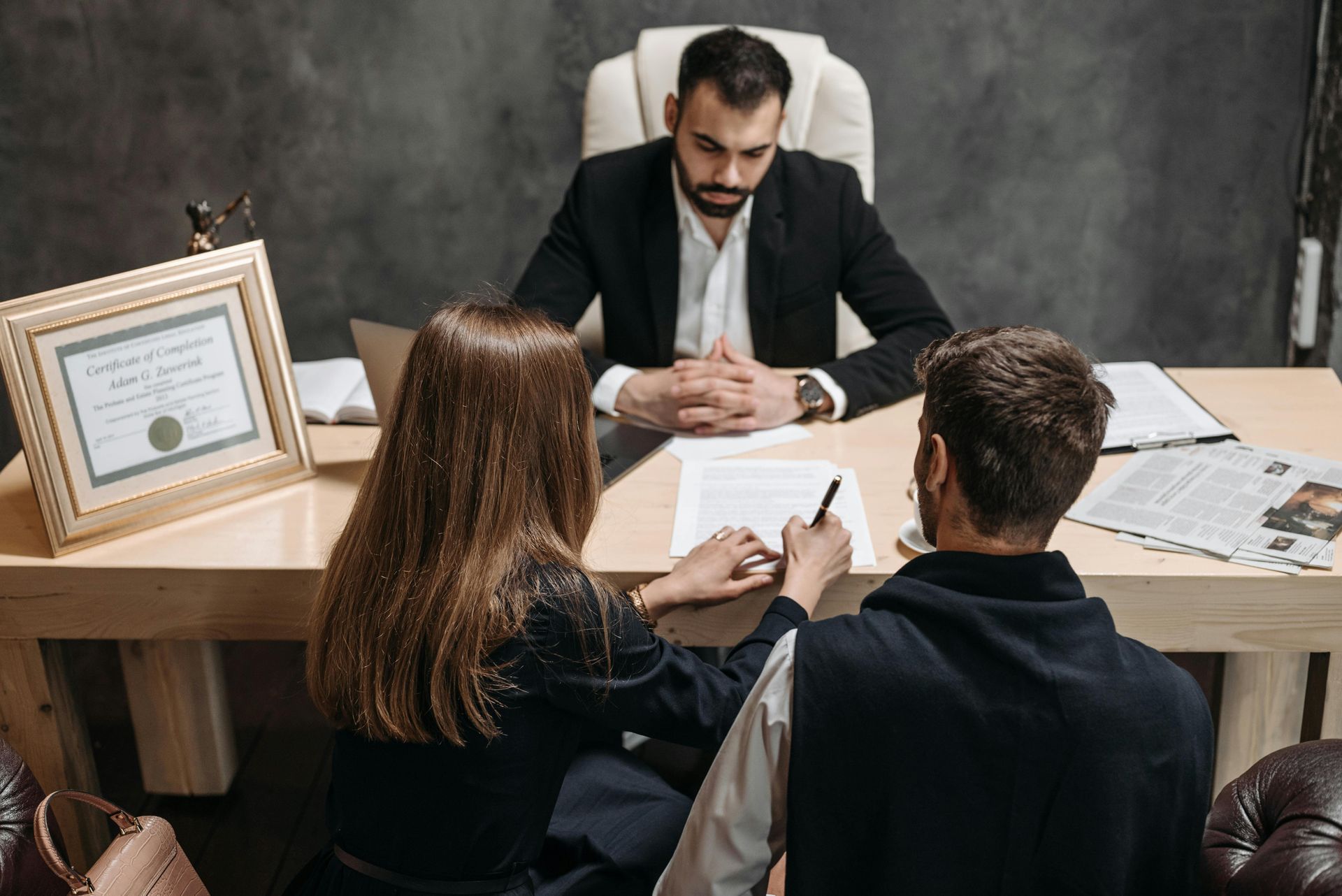 A couple signing documents at a desk while meeting with a professional in a formal setting.