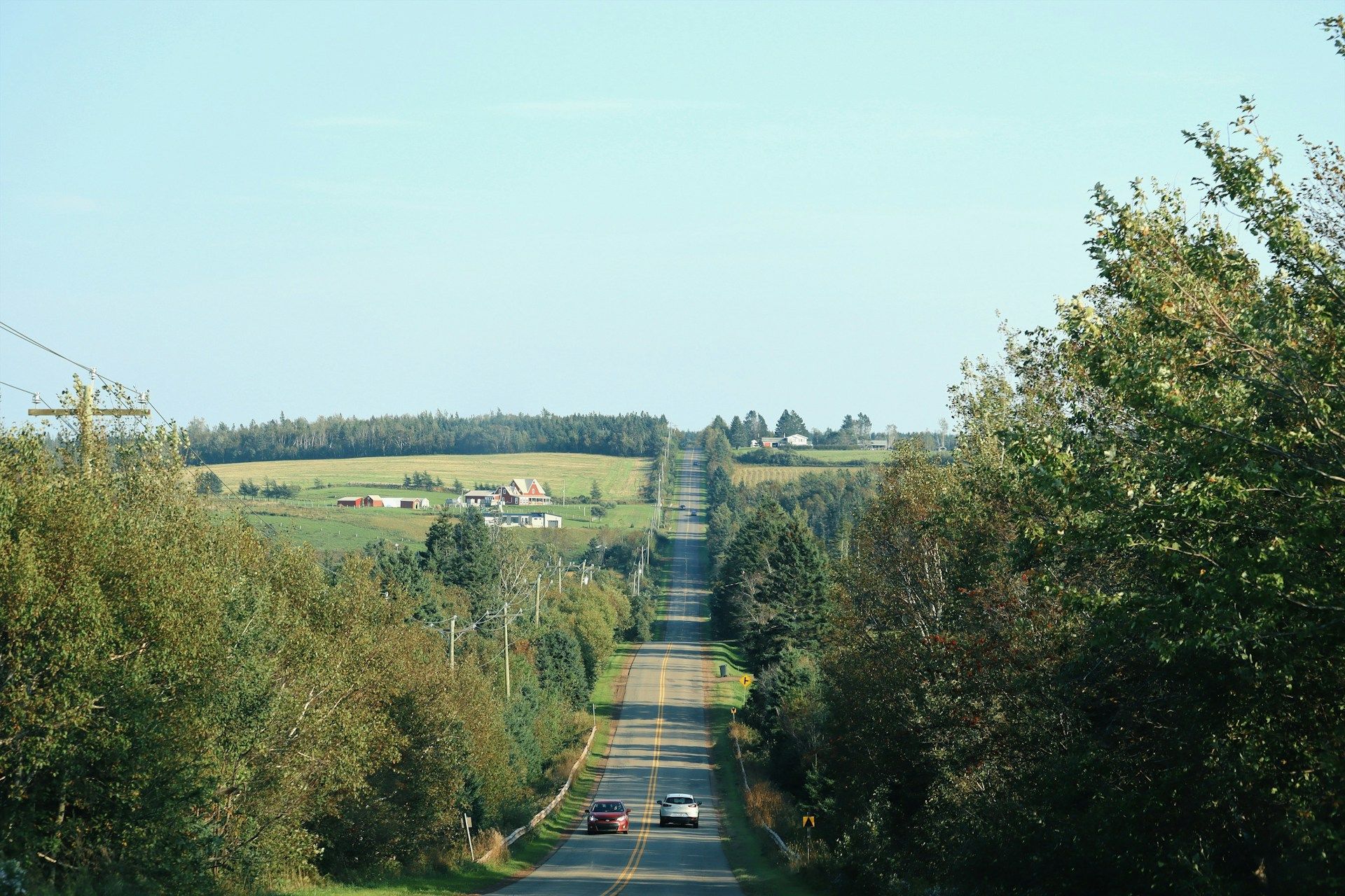 A long, straight road through a rural landscape, flanked by trees and fields under a blue sky. Two cars drive away.