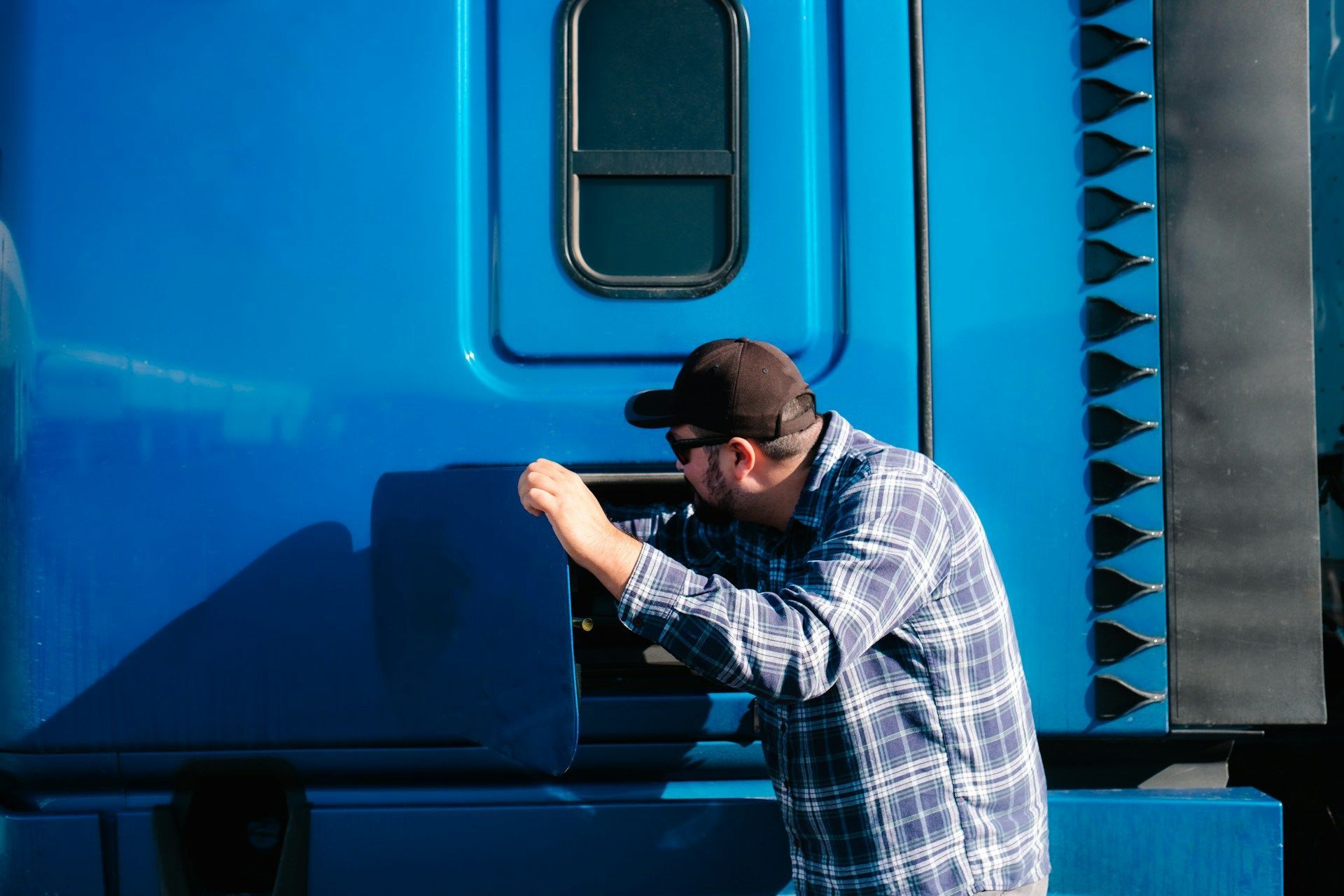 Man in plaid shirt and cap working on a blue semi-truck door outside.