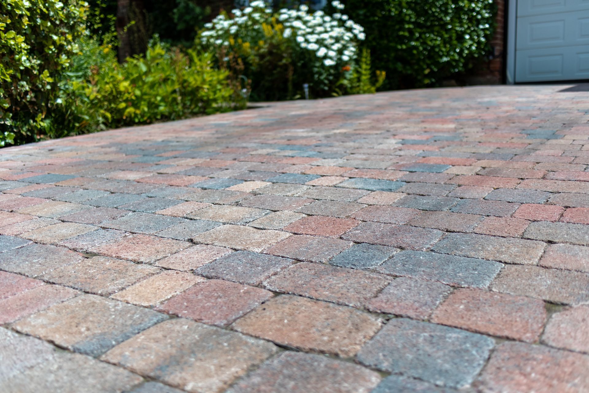 Colorful brick driveway leading to a garage, bordered by green shrubs and white flowers