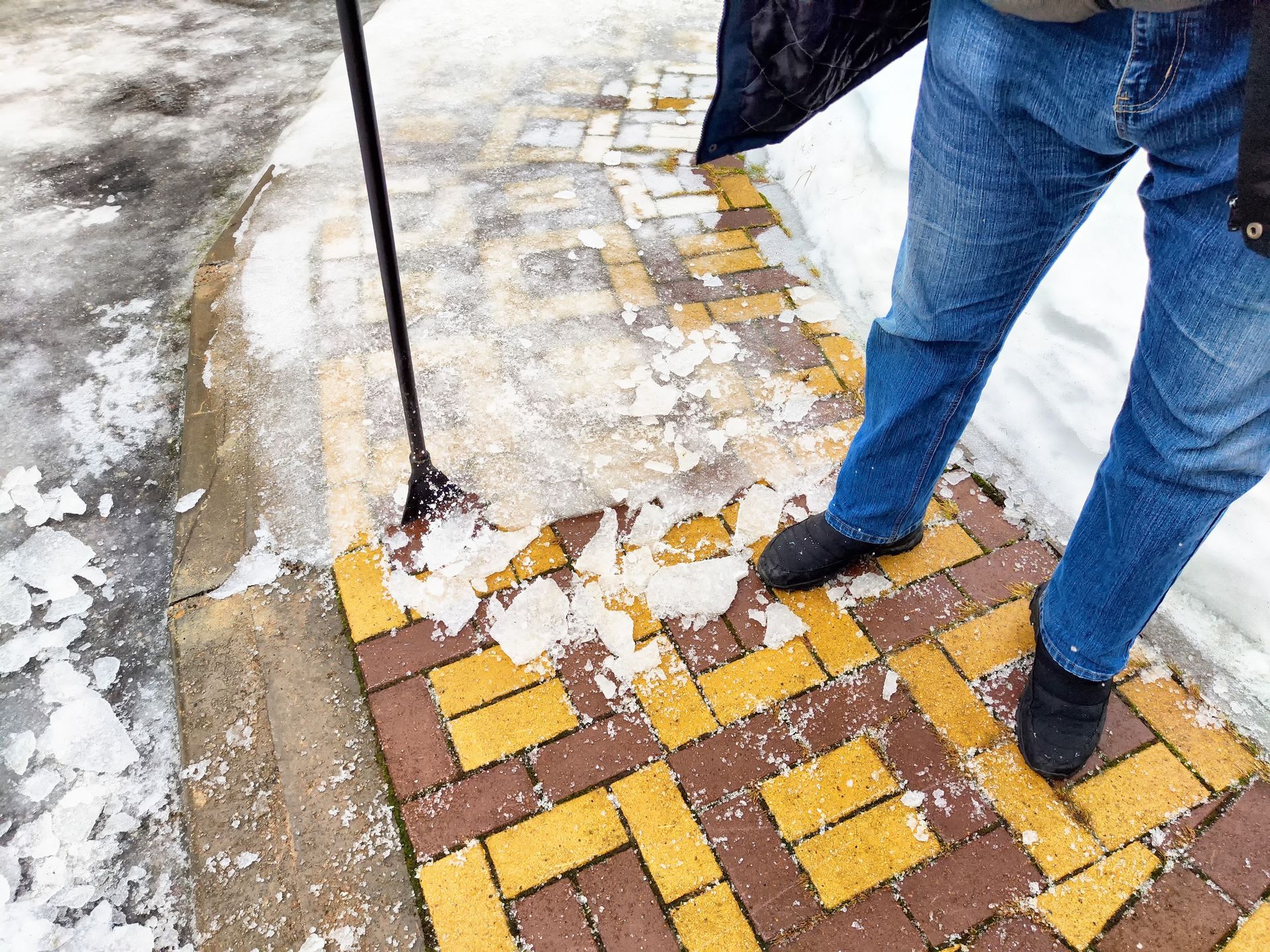 Person shoveling snow off a brick walkway, wearing blue jeans and black shoes.