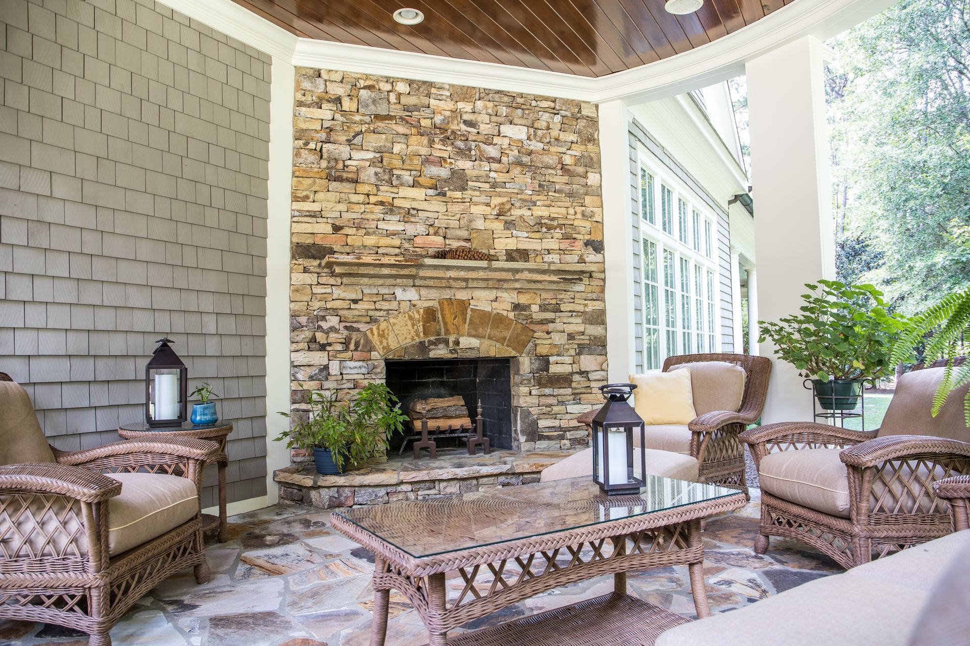 Covered stone patio with fireplace, wicker chairs, and glass coffee table with lanterns and potted plants