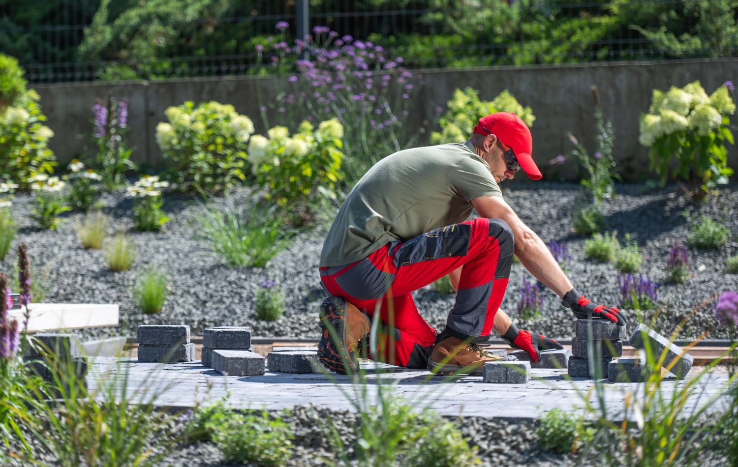 A gardener in red pants and cap kneels to lay pavers in a sunny flower bed.