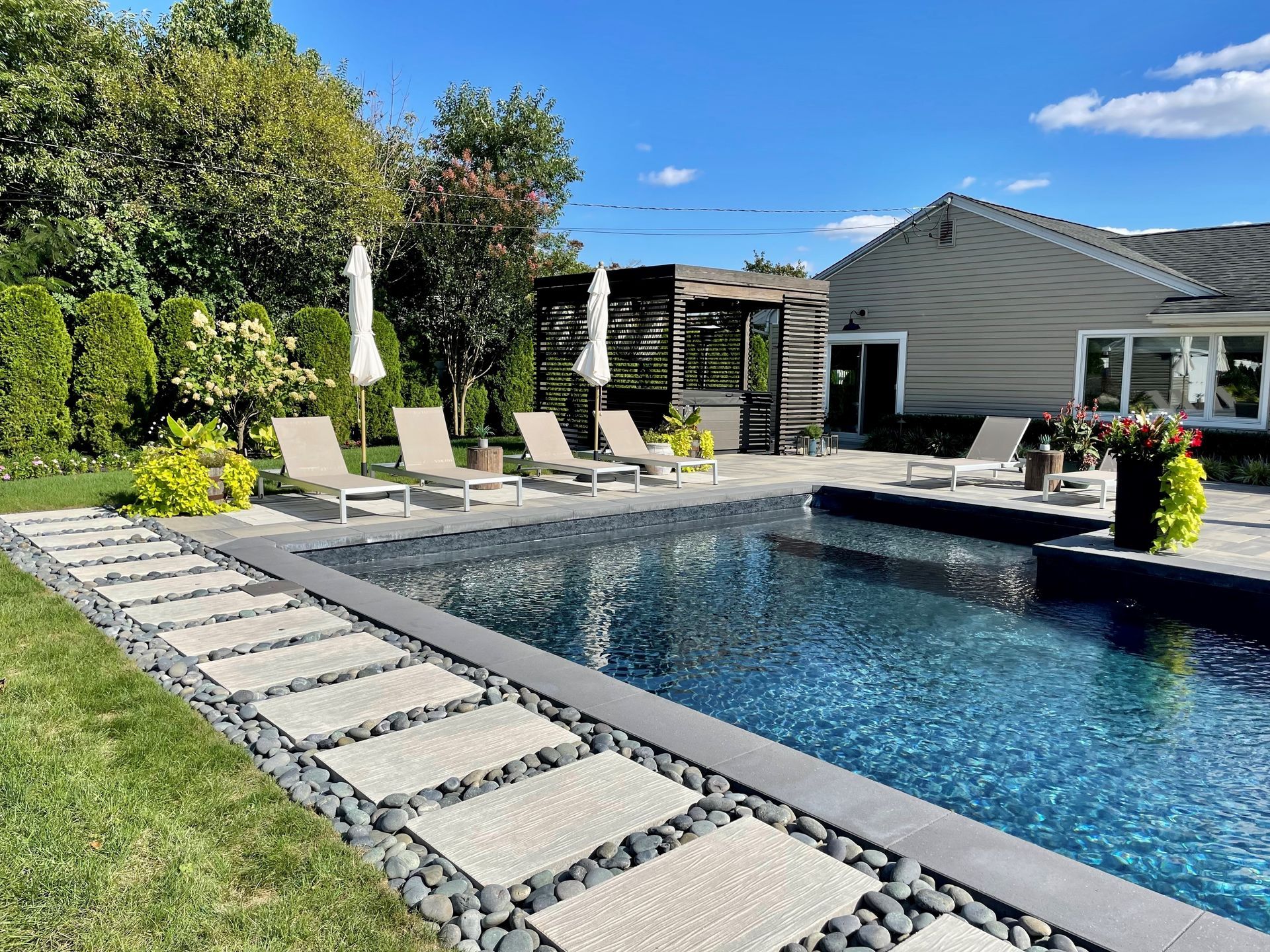 Backyard pool with lounge chairs, umbrella, and modern house under a sunny blue sky