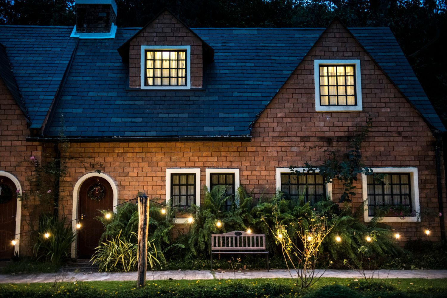 Brick house at dusk, lit windows, garden with fairy lights, bench.