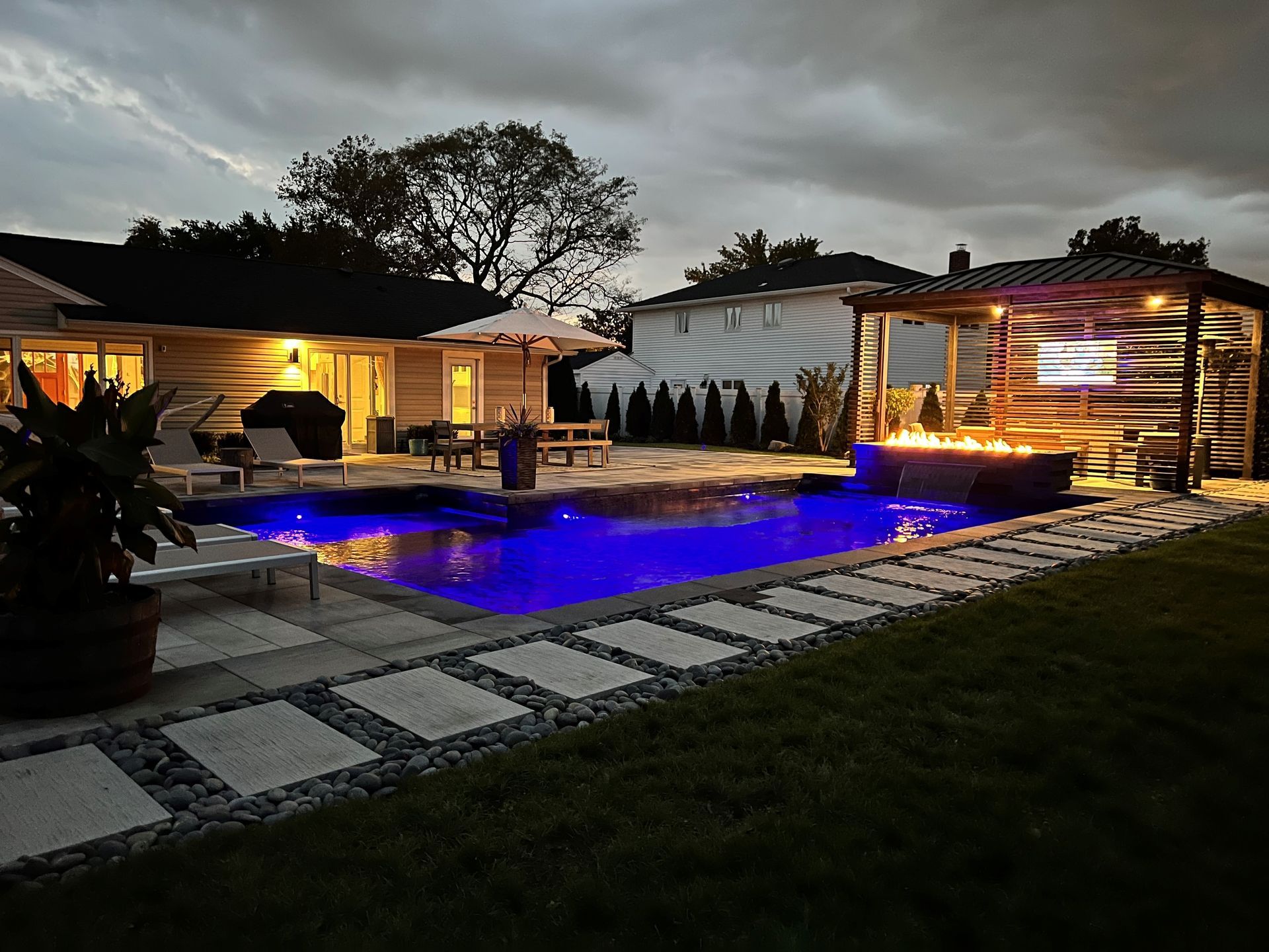Backyard pool at dusk lit with blue lights, with a fire pit and covered seating.