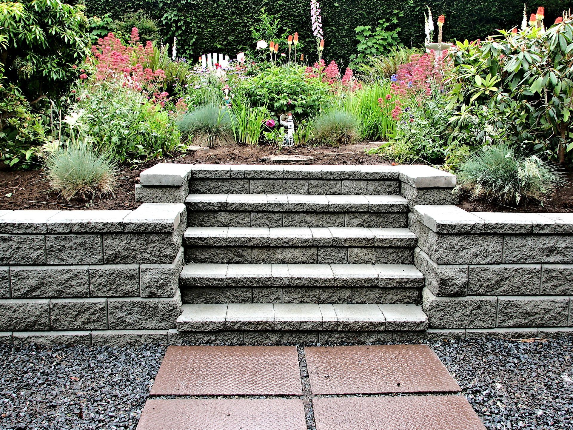 Stone steps leading to a lush garden with flowers and greenery, bordered by gravel and pavers